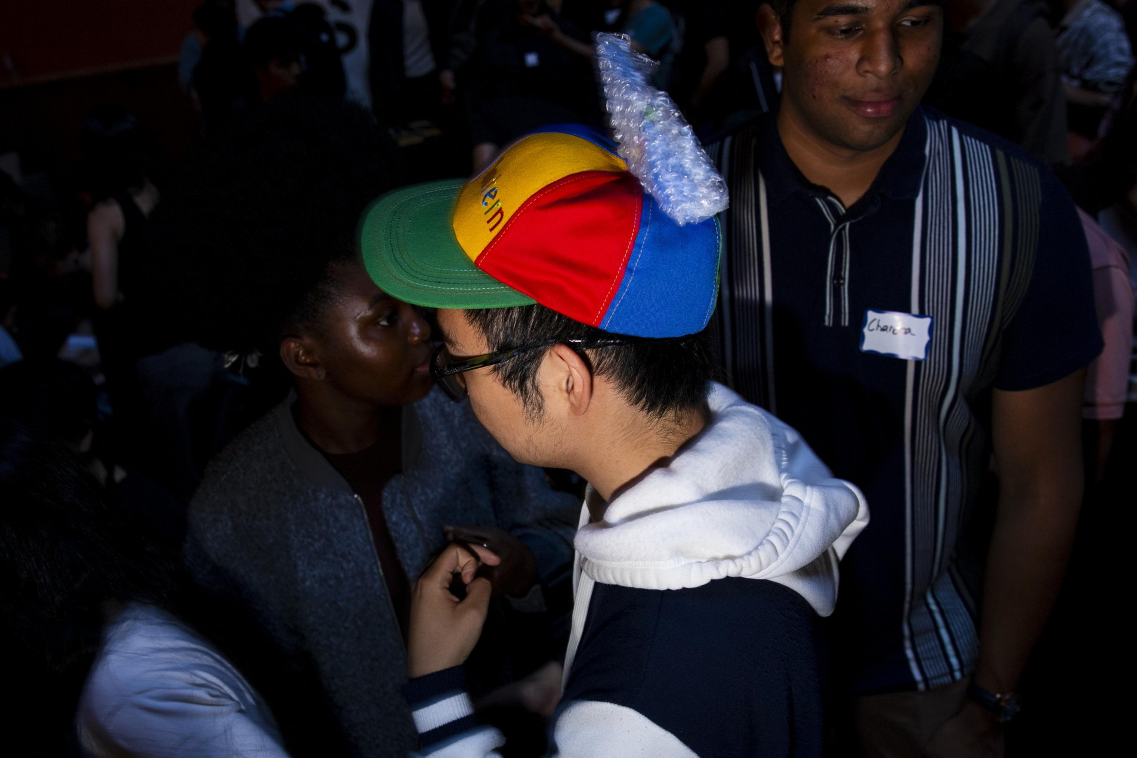 A person wearing glasses and a colorful propeller beanie interacts with others in a dimly lit crowded space, while a man with a name tag stands nearby.