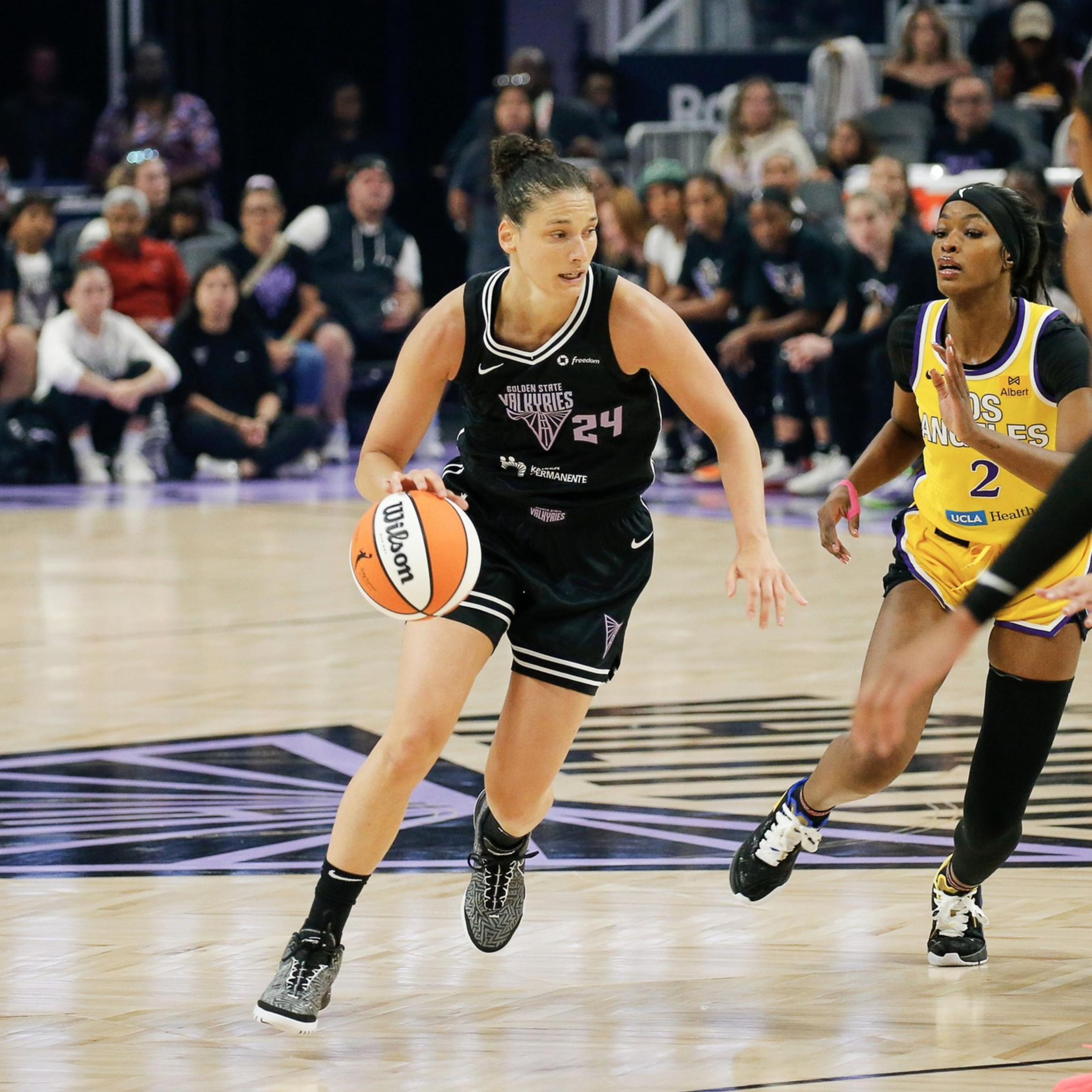 A basketball player in a black uniform dribbles past two defenders in yellow uniforms during an intense game with spectators watching.