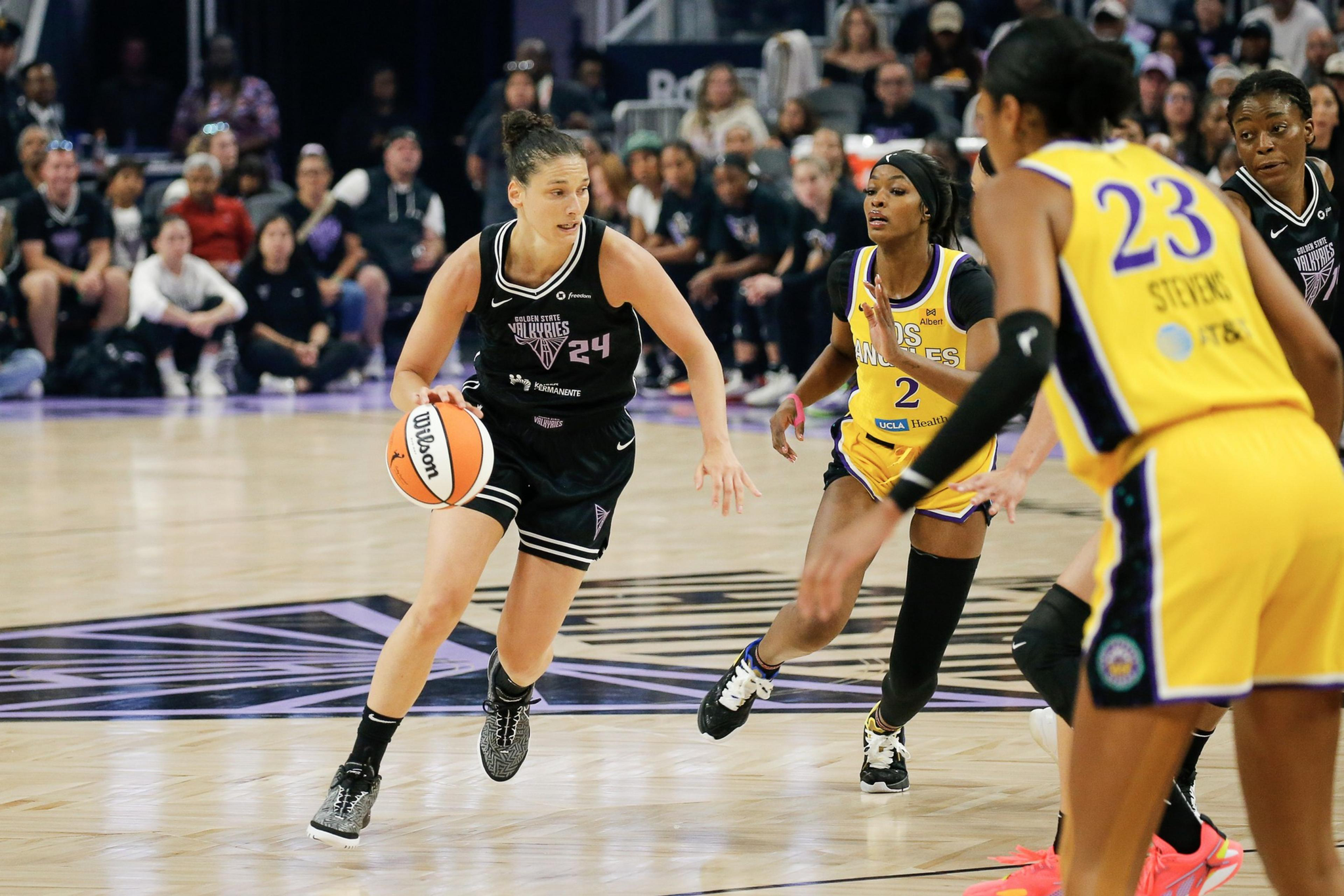 A basketball player in a black uniform dribbles past two defenders in yellow uniforms during an intense game with spectators watching.