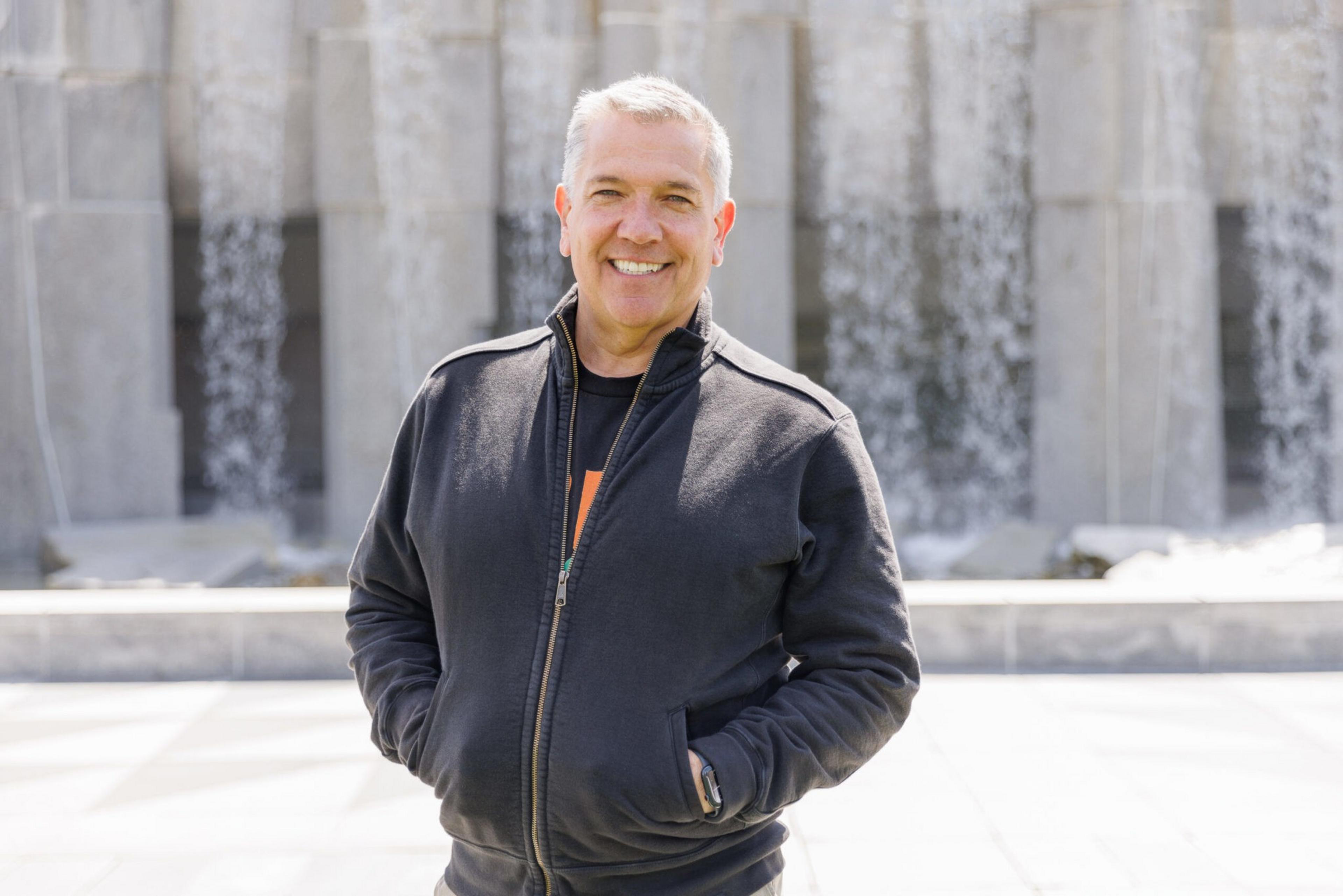 A smiling man in a black jacket stands in front of a wall with waterfalls.
