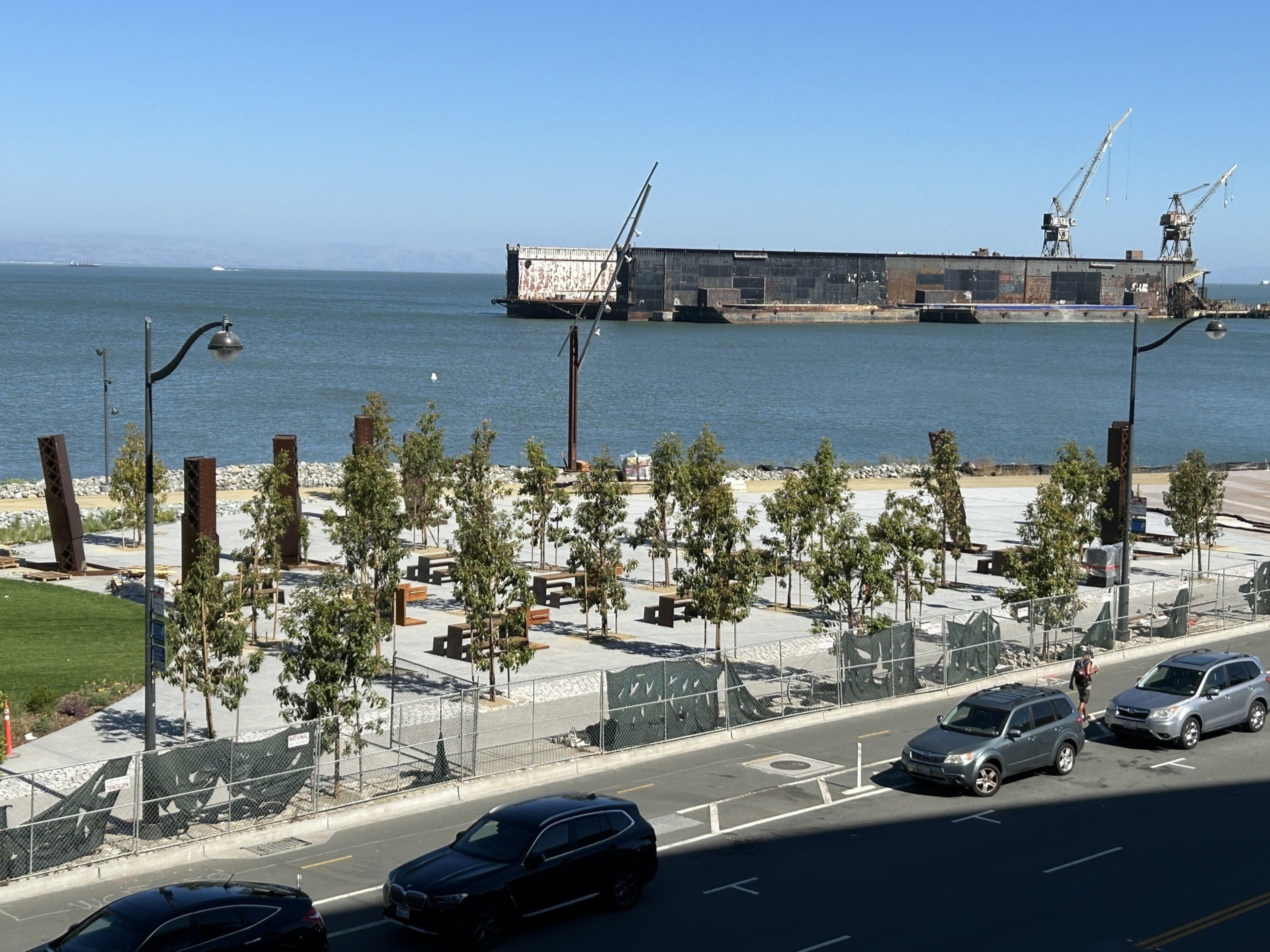 The image shows a waterfront area with a park filled with benches and trees. In the background, there's an industrial site with cranes and a large rusted ship.