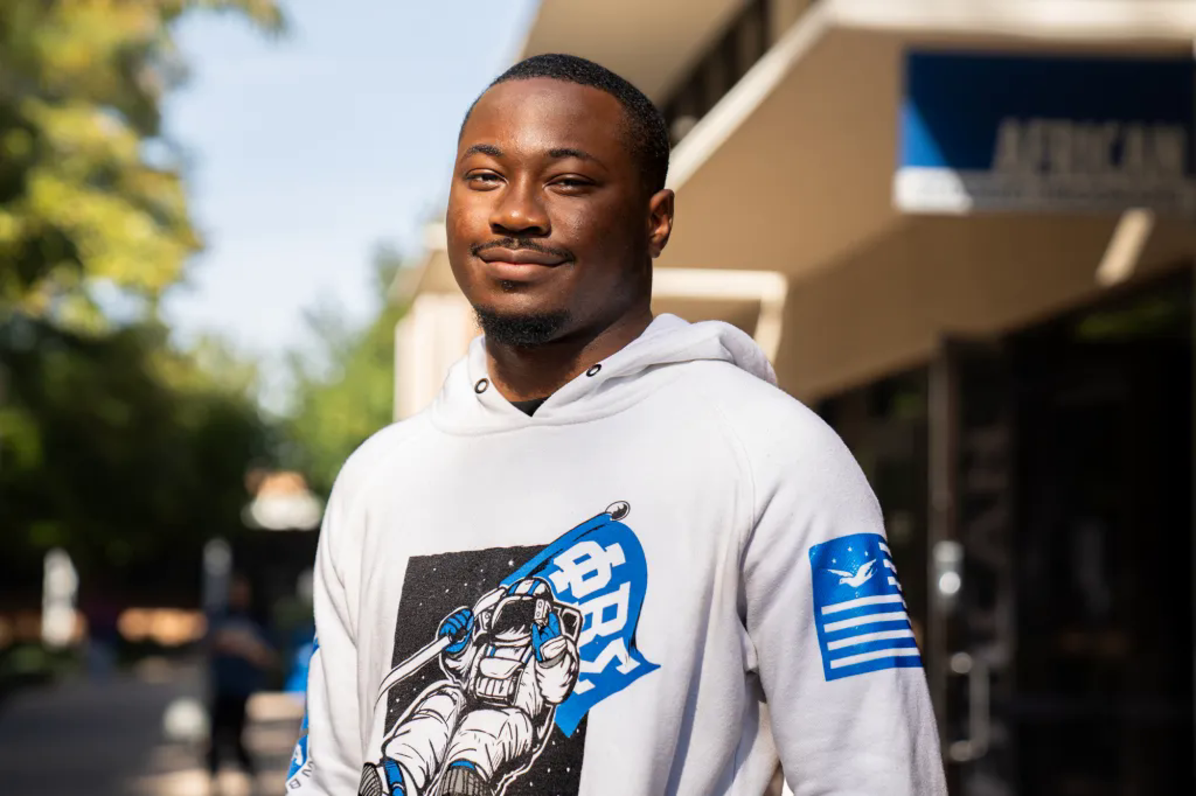 A content man in a white hoodie with a graphic print, outdoors on a sunny day.