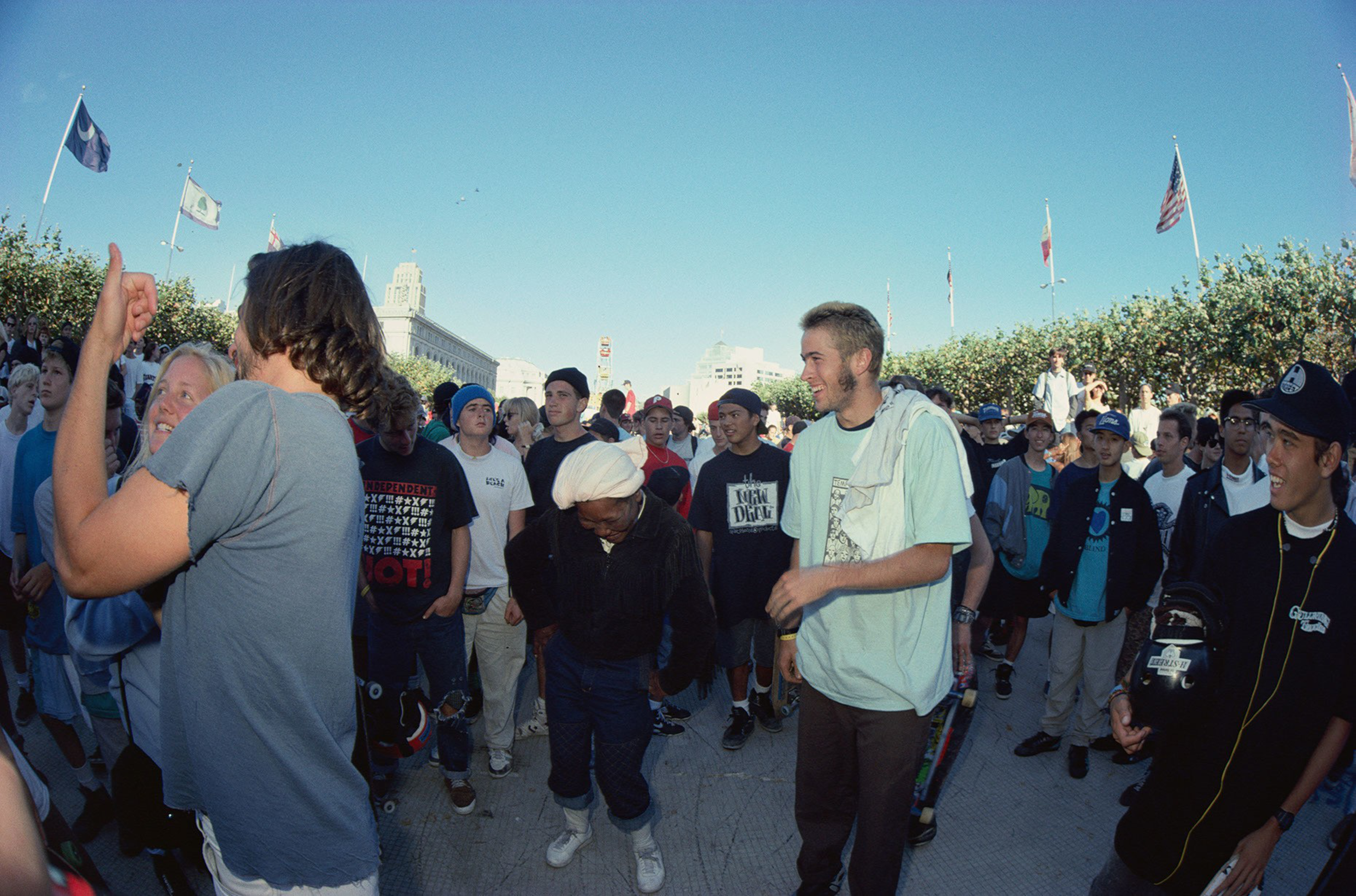 A diverse group of young people stand and socialize outdoors under a clear blue sky, some holding skateboards and wearing casual streetwear.