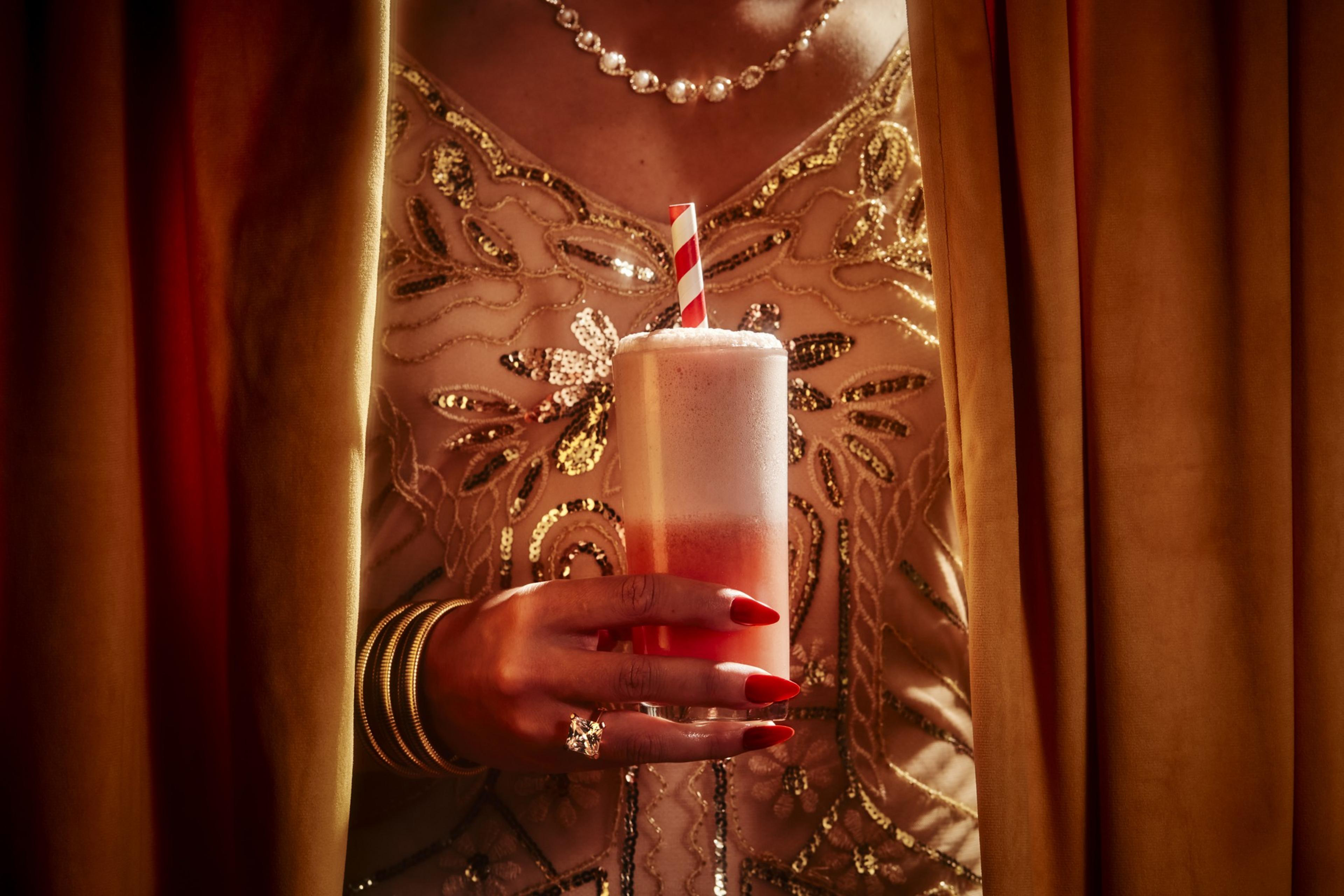 A woman in an ornate gold dress holds a pink drink with foam and a striped straw, partially hidden by gold curtains, showcasing jewelry on her hand.