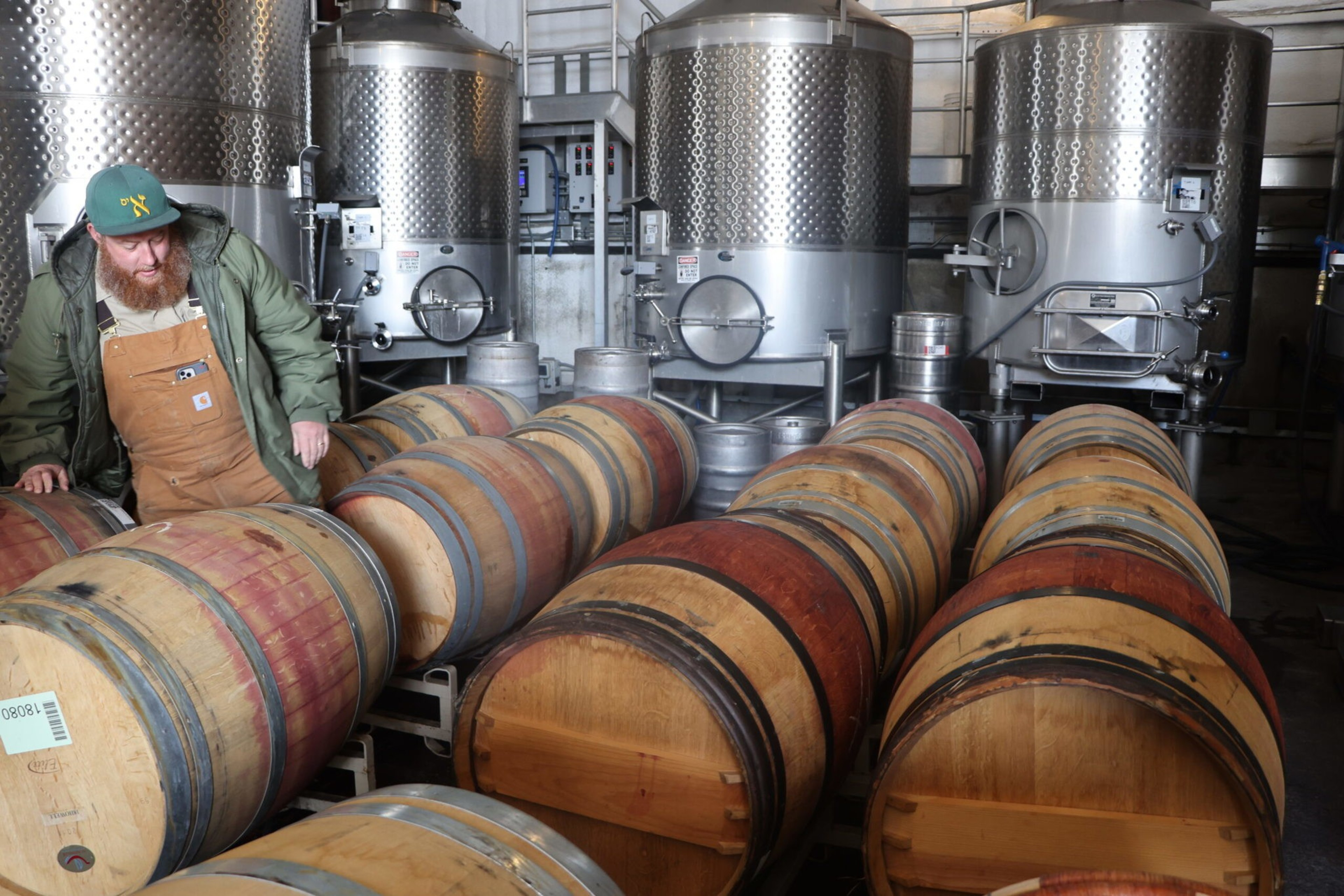 A man in a green jacket and brown overalls inspects wooden barrels stacked horizontally in front of large stainless steel tanks.