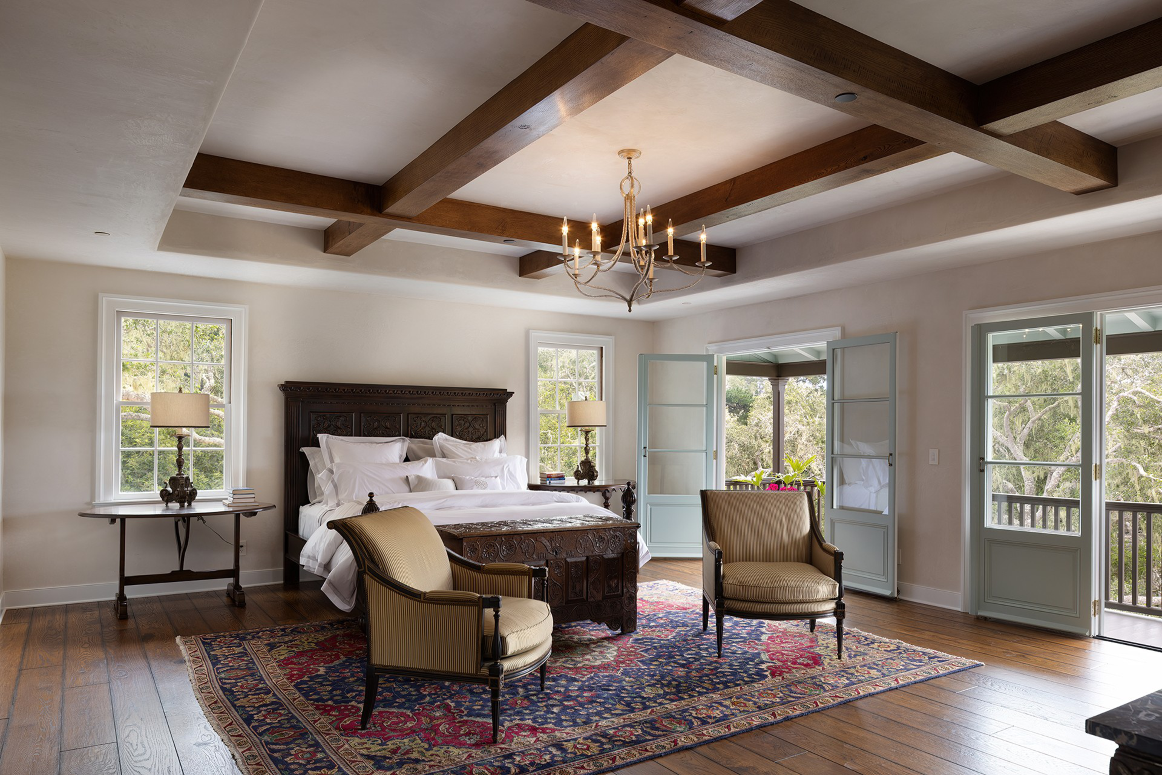 A spacious bedroom with a carved dark wood bed, two armchairs, a colorful rug, wooden beams on the ceiling, and open doors leading to a balcony.
