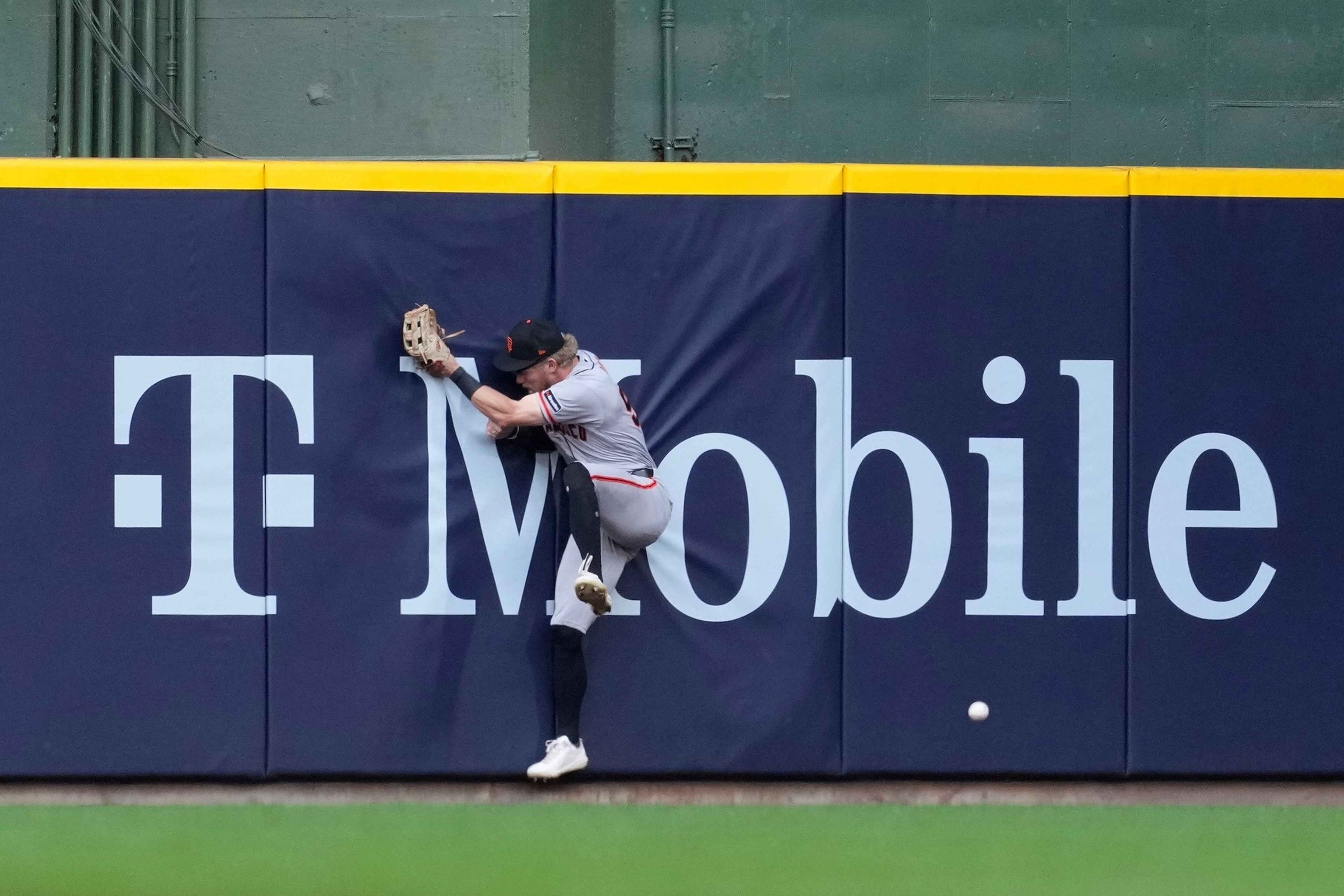 A baseball player in grey uniform jumps against a padded outfield wall with a glove raised, as a baseball rolls on the grass nearby.
