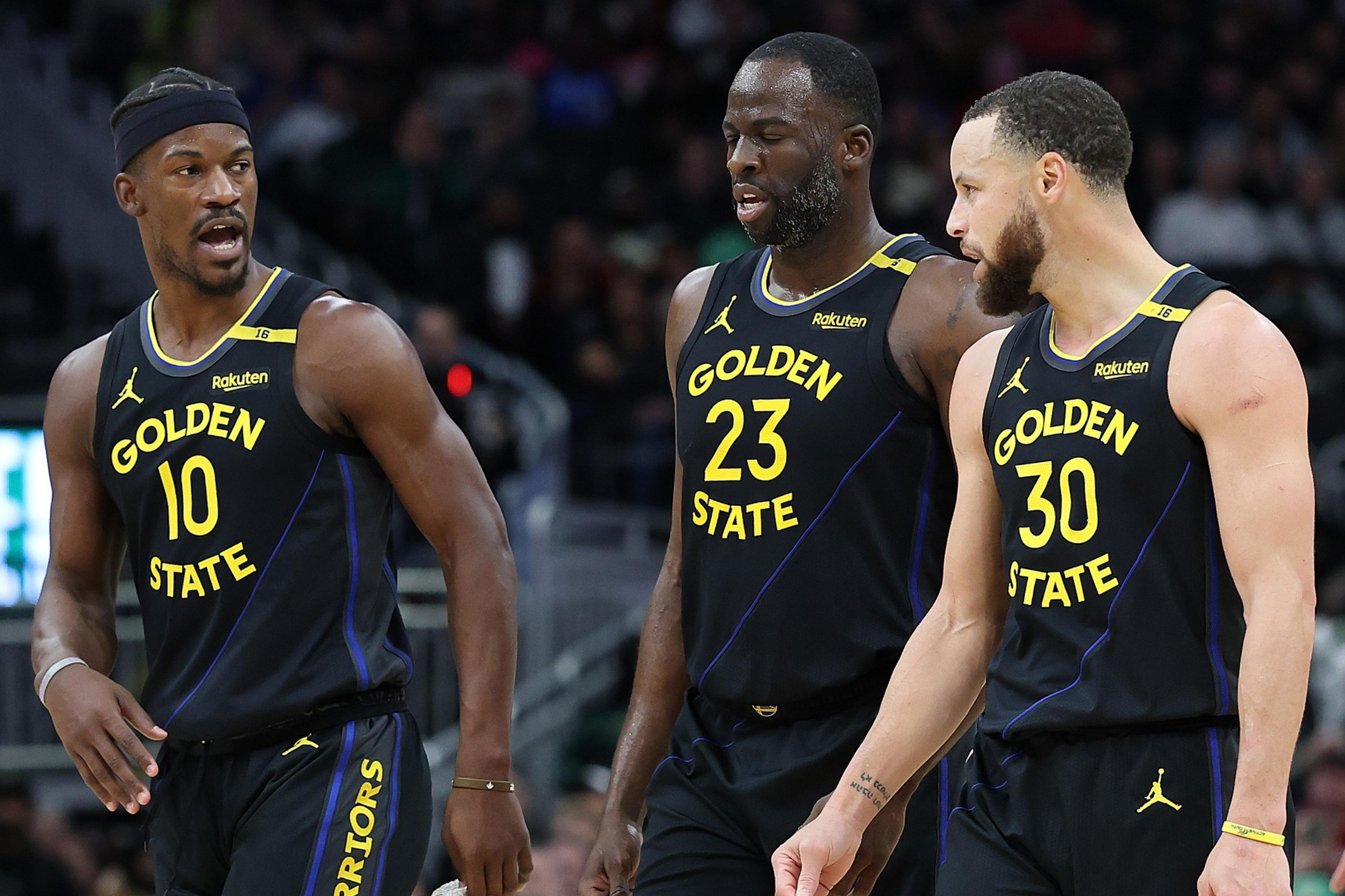 Three basketball players in black "Golden State" uniforms converse on the court, with one gesturing animatedly while walking.