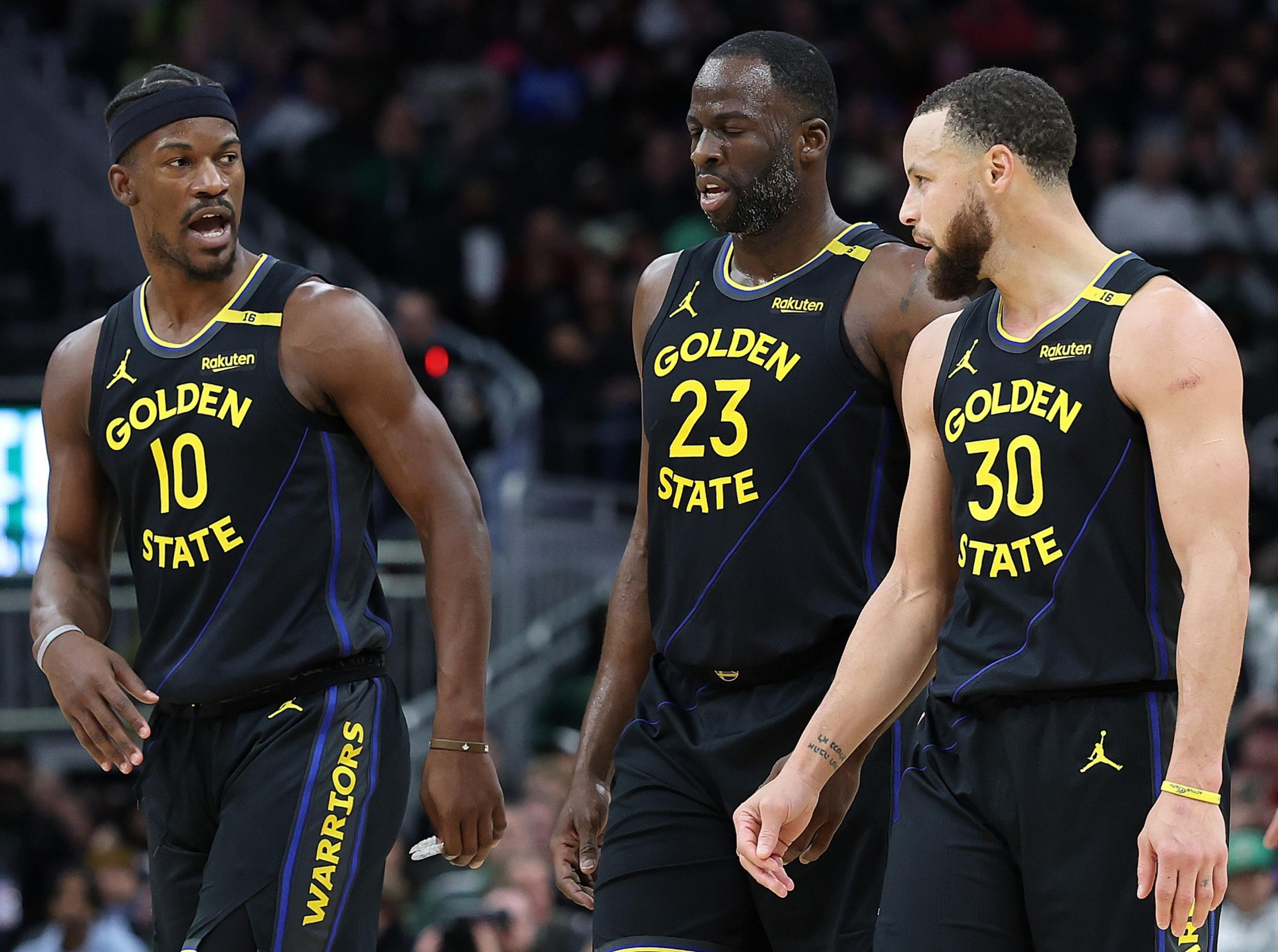 Three basketball players in black &quot;Golden State&quot; uniforms converse on the court, with one gesturing animatedly while walking.