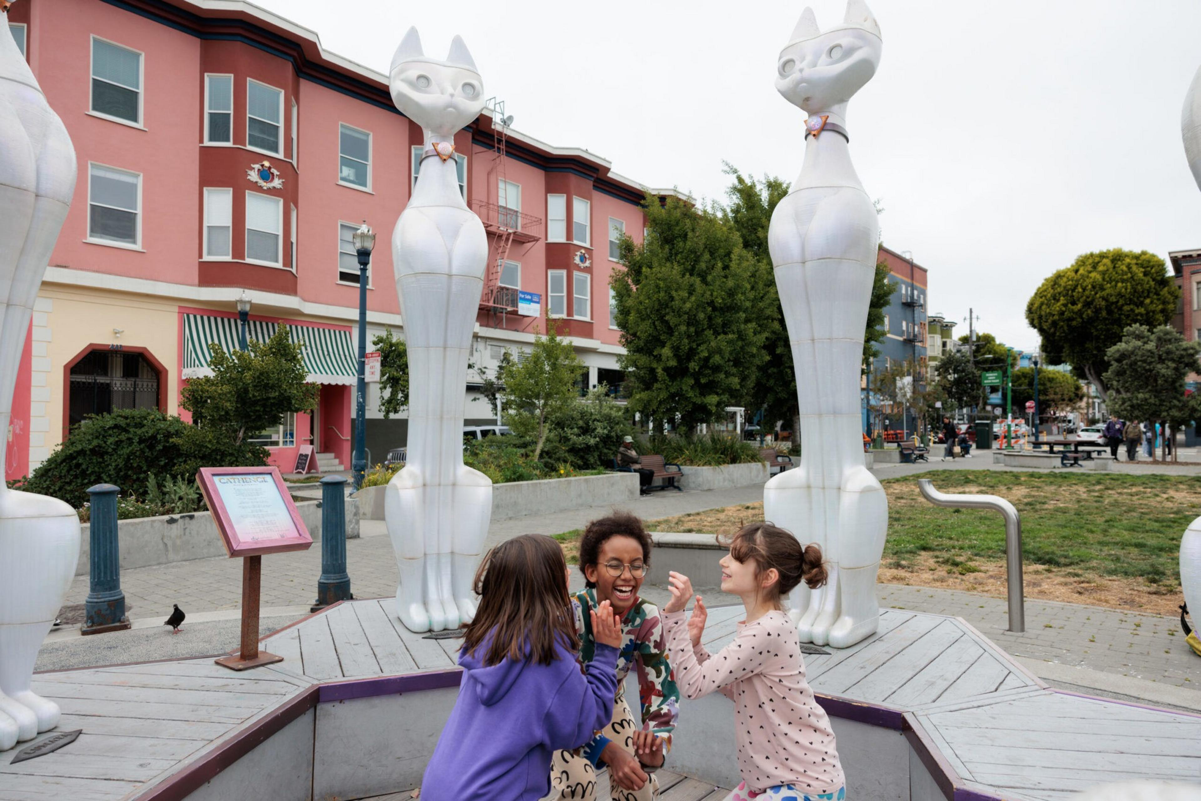 Three children laugh and play near tall white cat sculptures in an urban park with buildings, trees, and benches in the background.