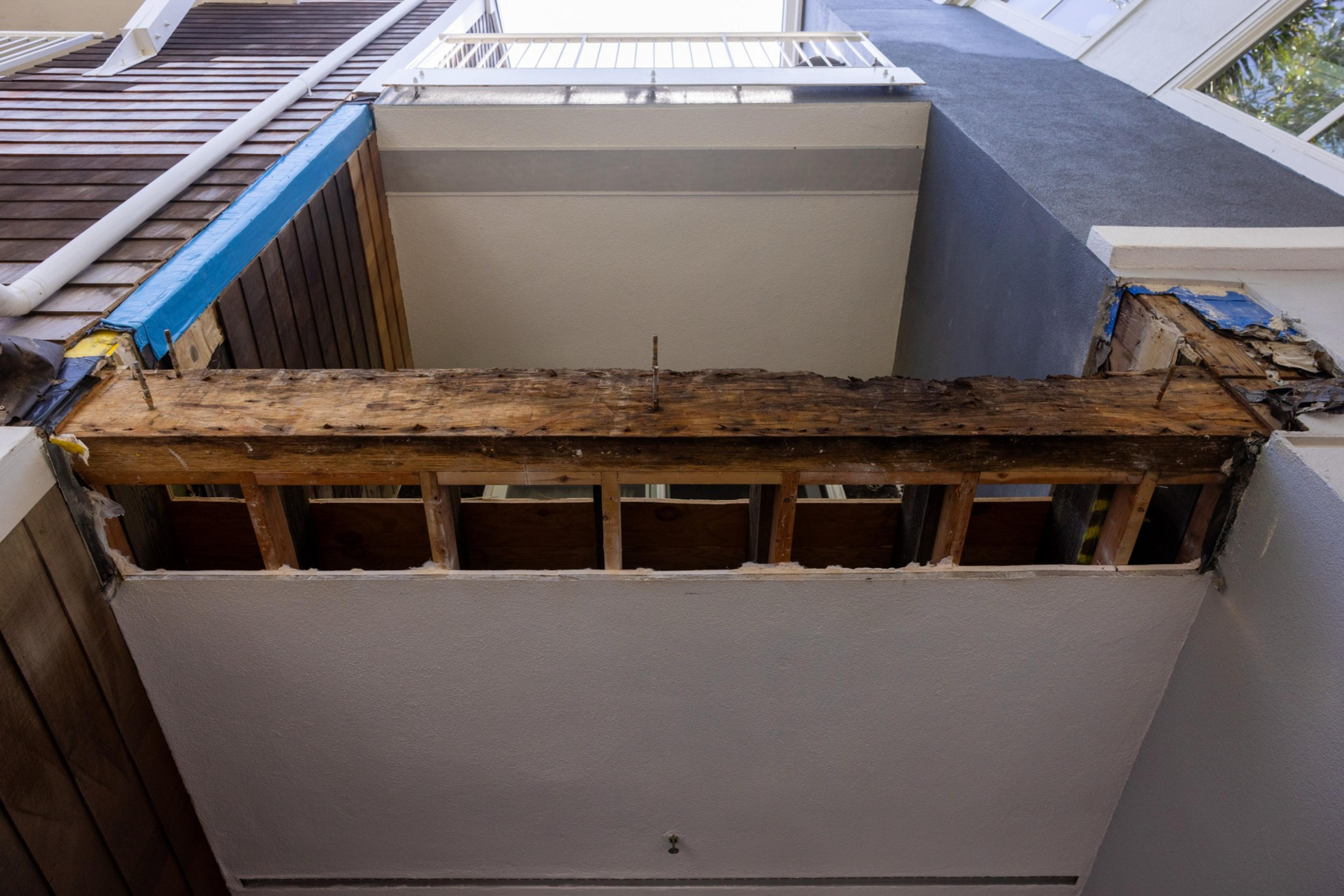 The image shows a partially demolished wooden beam structure under a small open area, framed by walls, with visible blue tape and exposed wood.