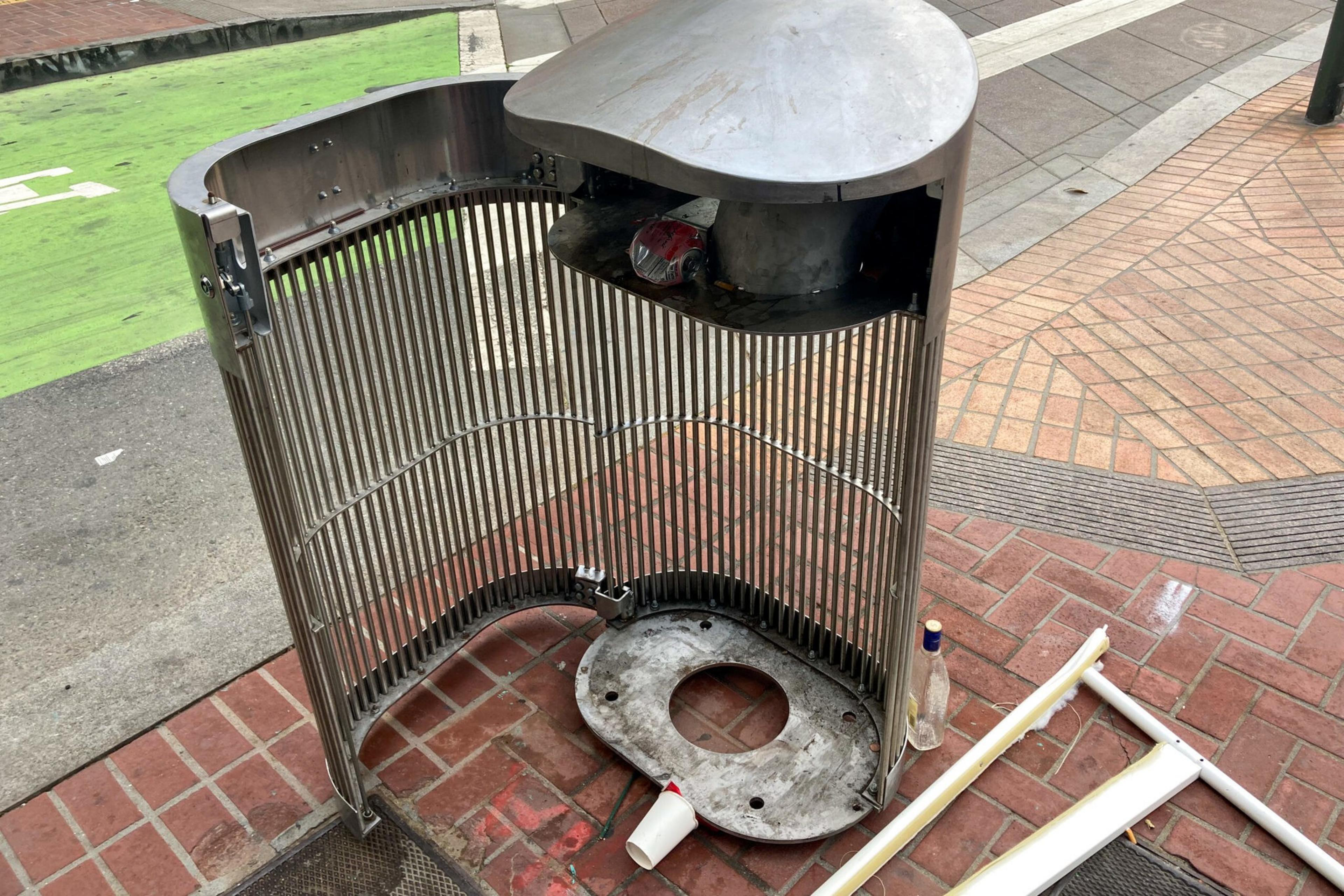 A dismantled, metal public trash can on a red brick sidewalk; the lid and base are removed, leaving only the frame. Debris and a bottle lie nearby.