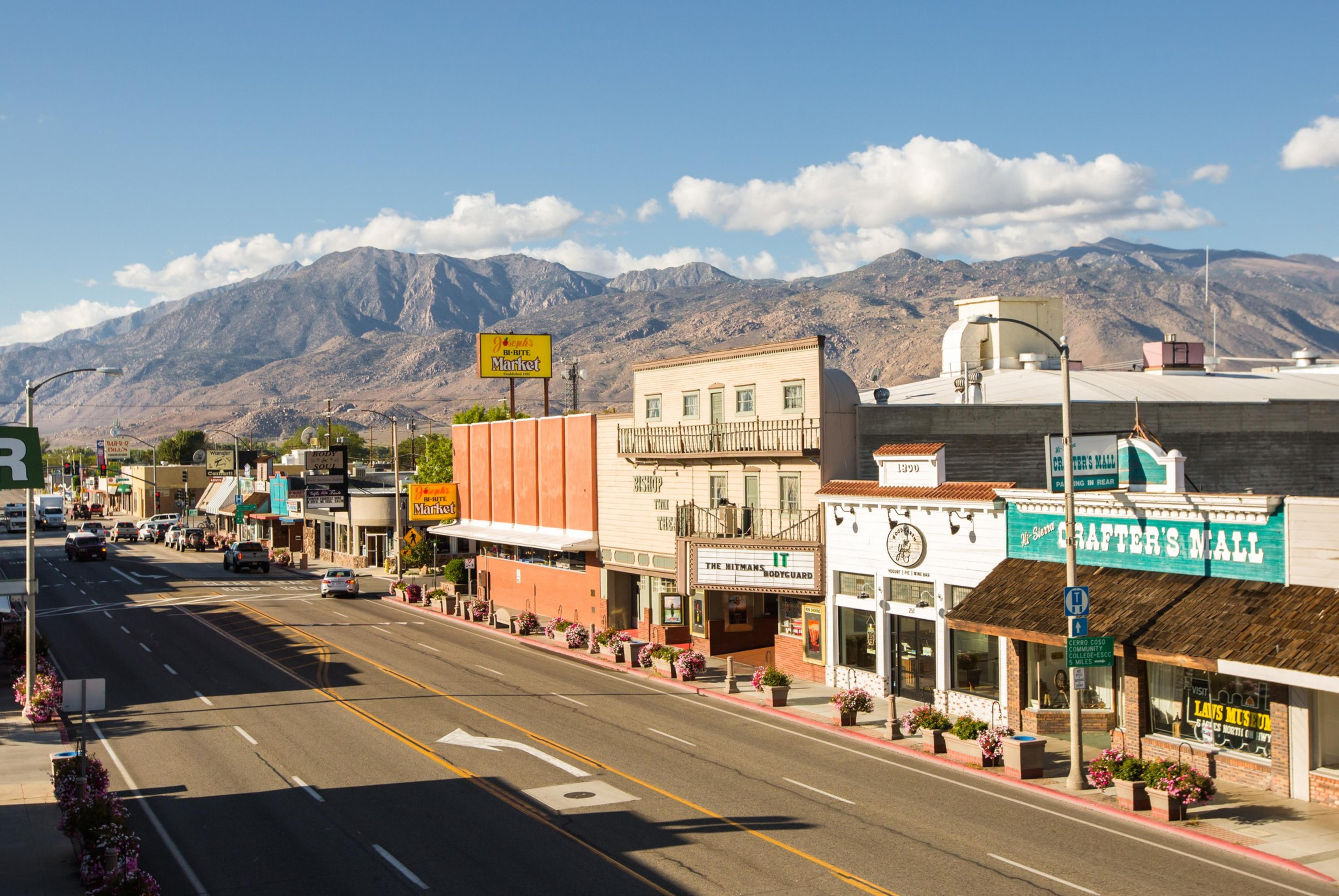 A small town's main street with low-rise buildings, shops, and signs, set against a backdrop of rugged mountains under a blue sky with scattered clouds.