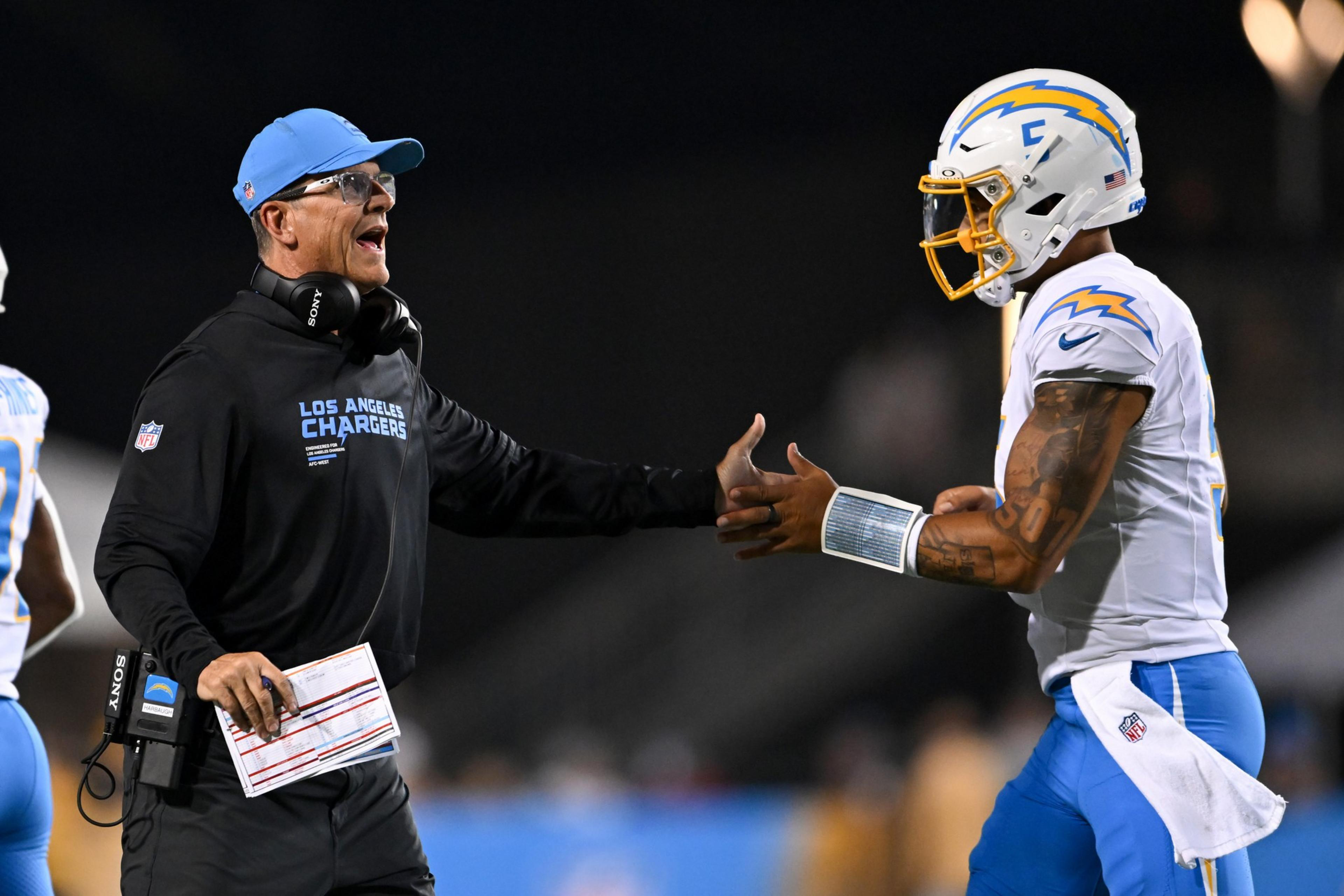 A coach wearing a black Los Angeles Chargers outfit and blue cap reaches to high-five a football player in white and blue Chargers gear and helmet.