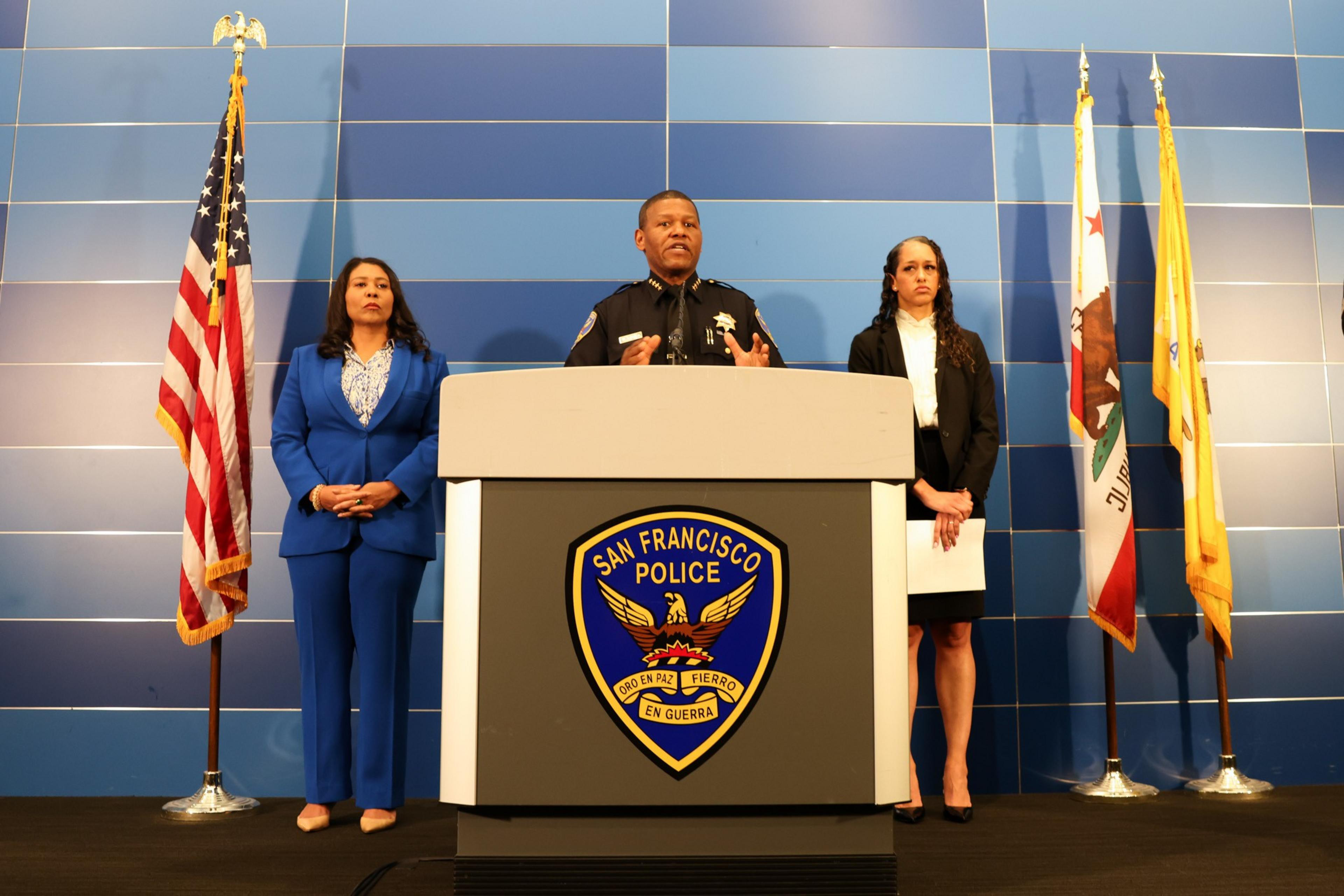 Three people stand at a podium with the San Francisco Police logo, flanked by the US and California flags on a blue-background stage.