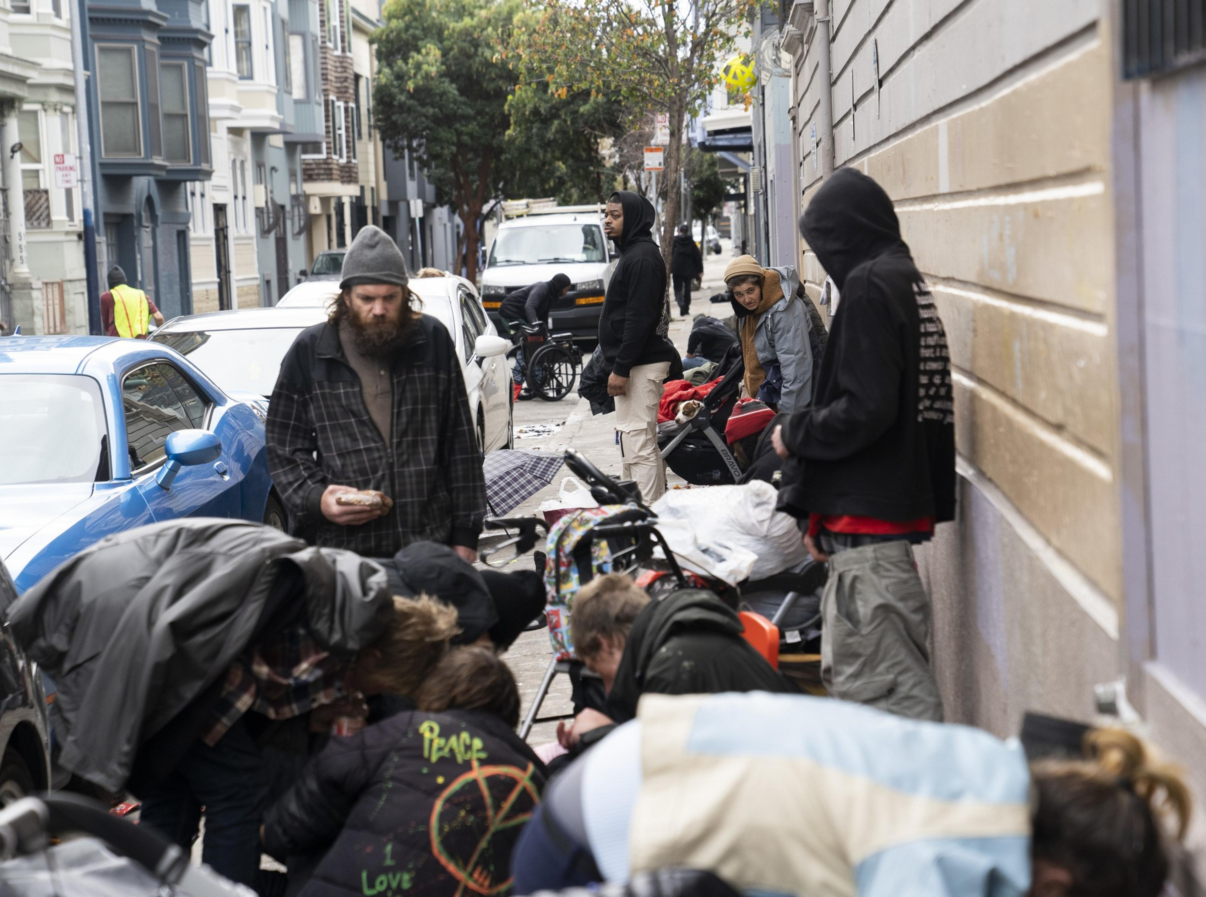 People stand and sit on a sidewalk cluttered with belongings and strollers, beside parked cars and buildings, creating a crowded and busy street scene.