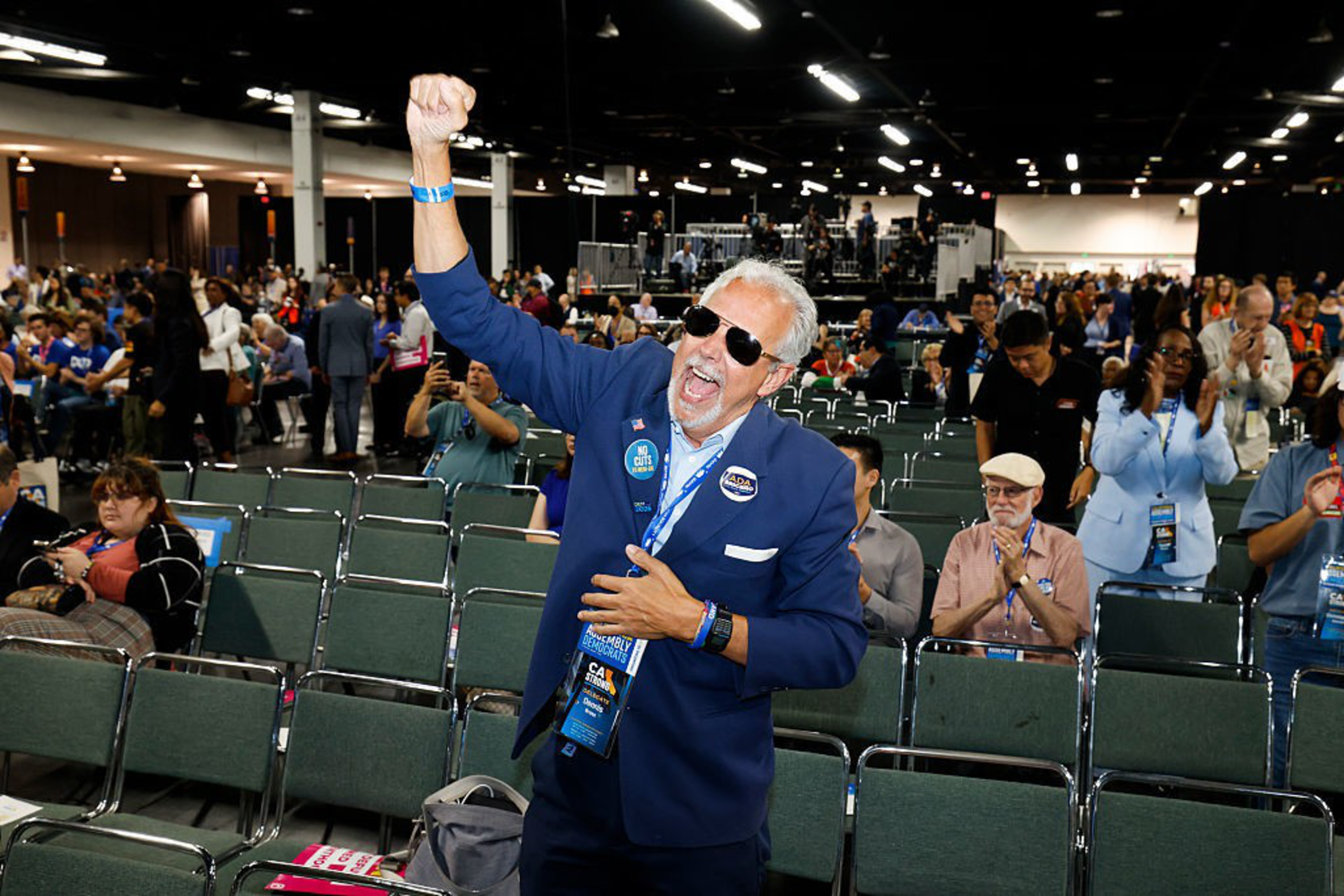 A man in a blue suit and sunglasses enthusiastically raises his arm, surrounded by a seated crowd in a large indoor event space.