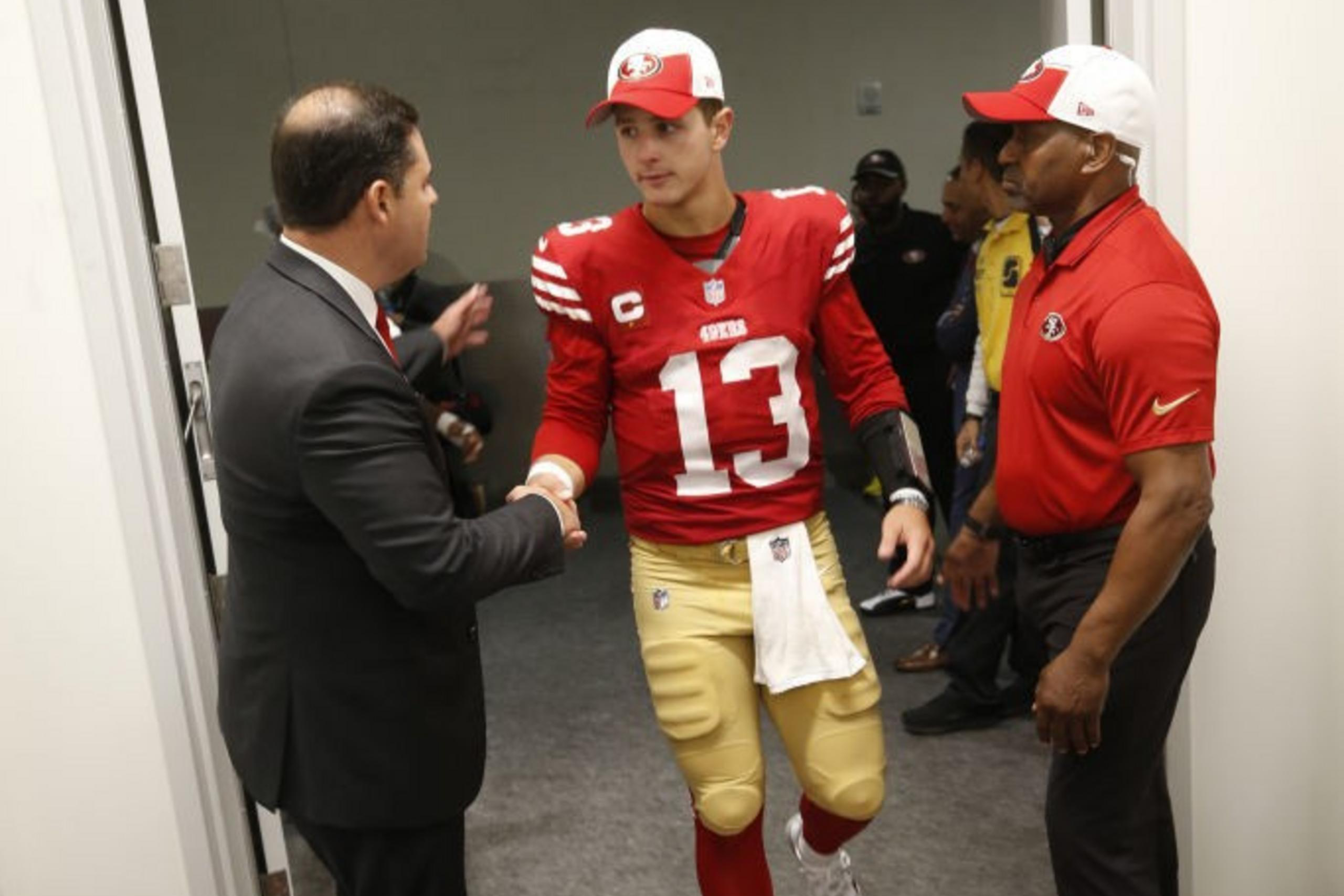 A football player in a red and gold uniform shakes hands with a man in a suit. Another man in a red shirt stands nearby. They are in a hallway.