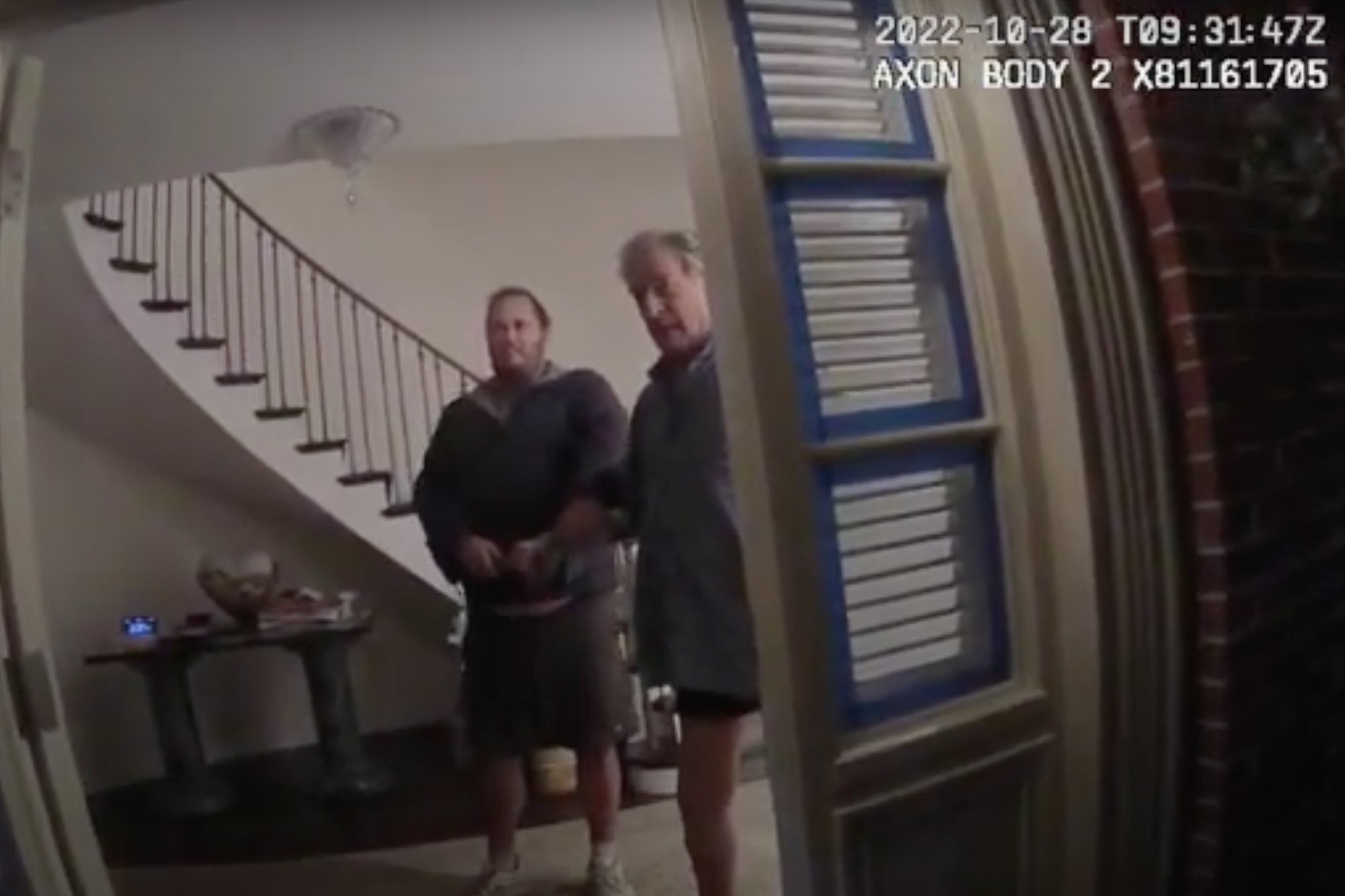 Two men, one wearing shorts and the other in a jacket, stand inside a house by the front door, which is ajar. A curved staircase is visible in the background.
