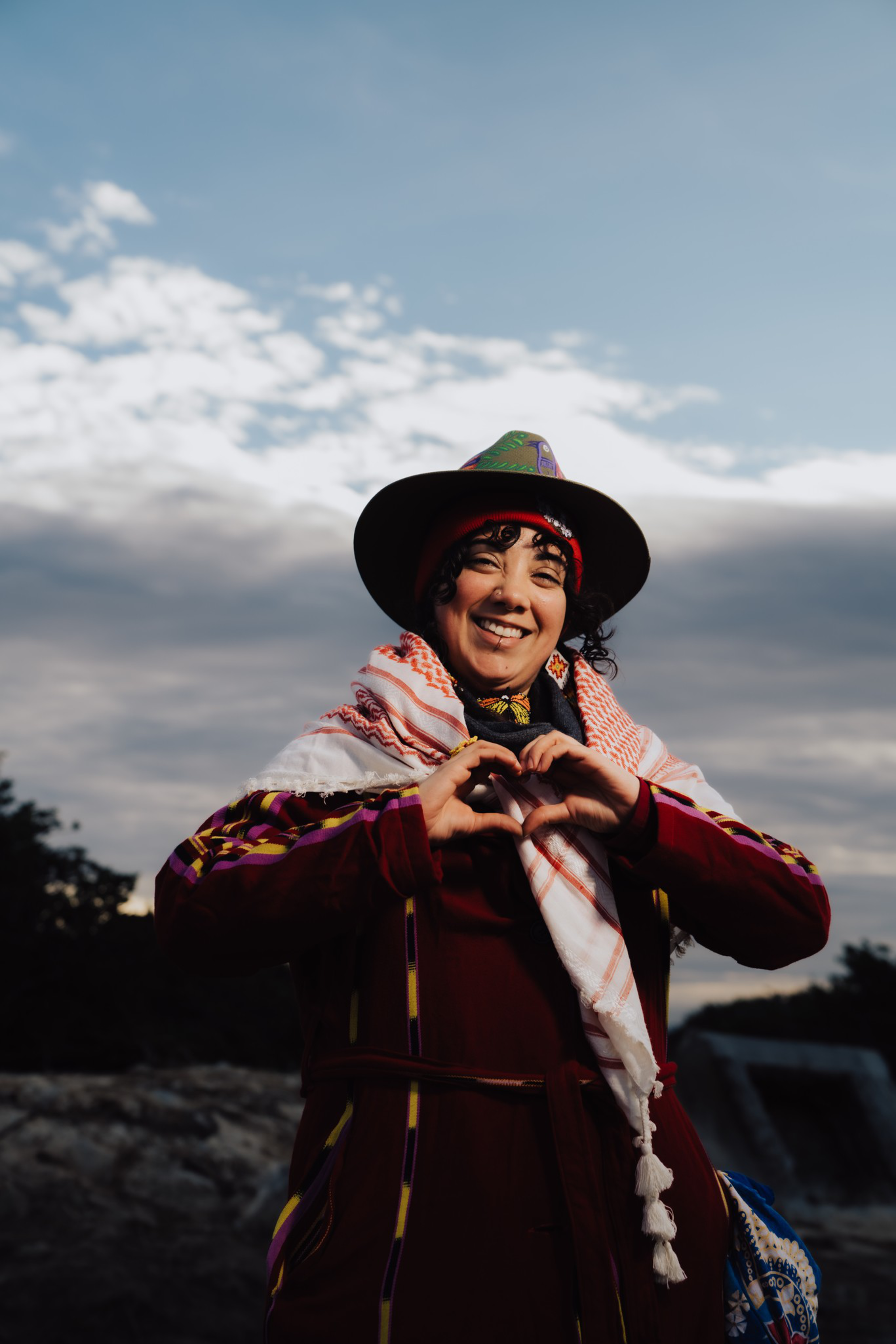 A smiling person wearing a hat, patterned scarf, and maroon jacket forms a heart shape with their hands against a cloudy sky.