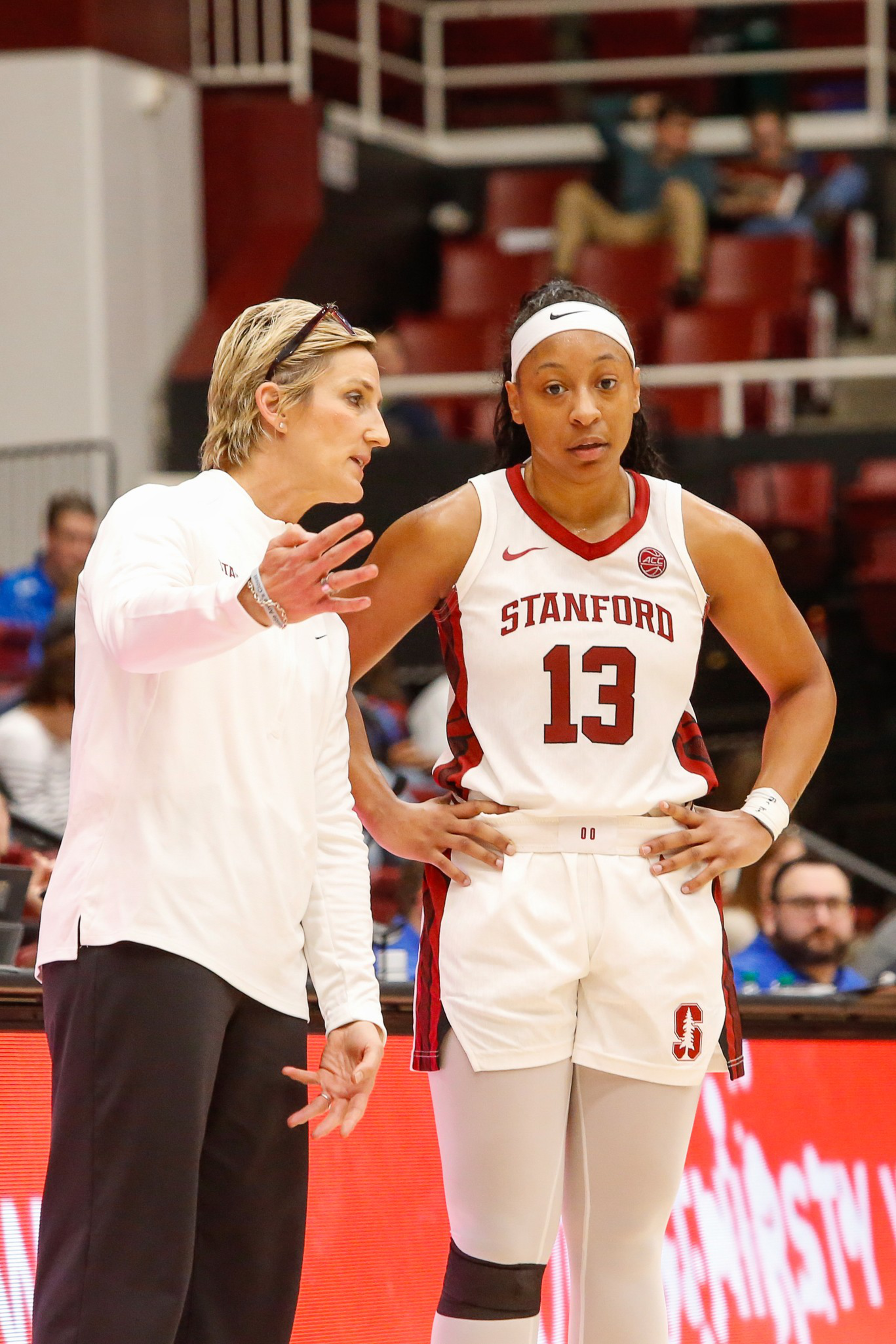 A coach in white passionately talks to a Stanford basketball player wearing jersey number 13, who listens with hands on hips during a game break.