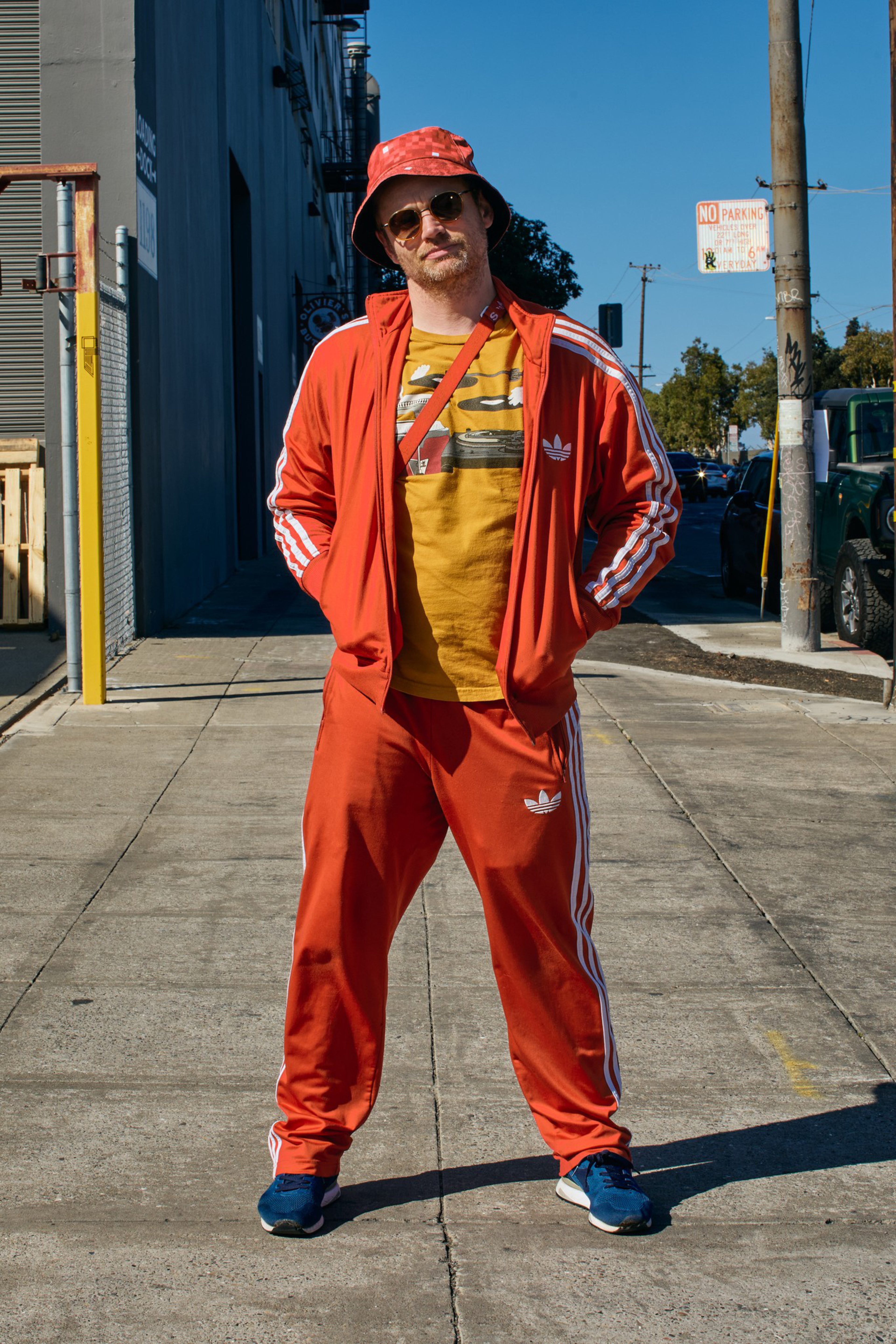 A man stands on a sunny sidewalk wearing an orange Adidas tracksuit, yellow graphic T-shirt, red bucket hat, sunglasses, and blue sneakers.