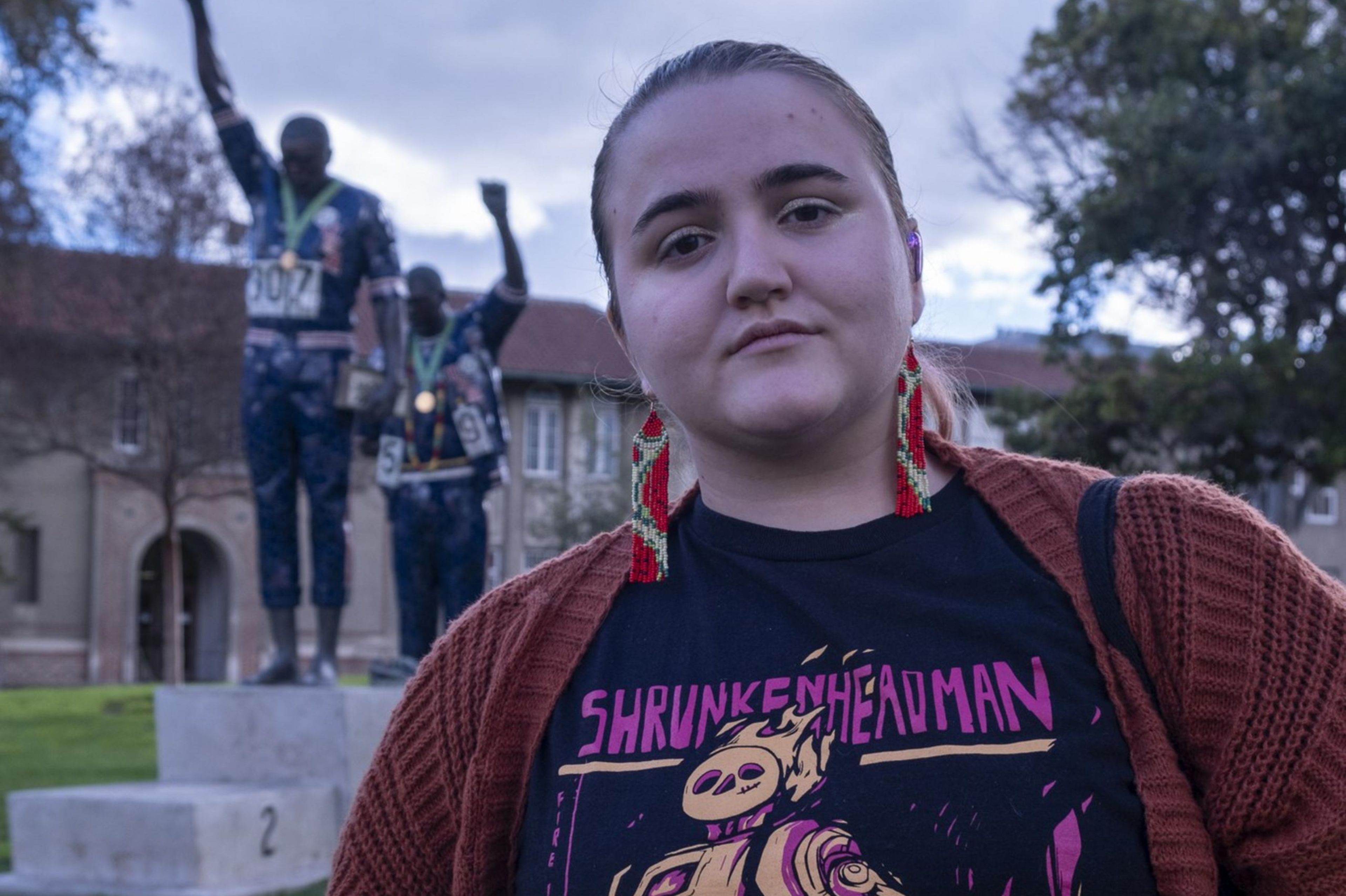 A person stands in the foreground wearing a graphic t-shirt and red cardigan. Behind them is a statue of two figures with raised fists, set against a cloudy sky.