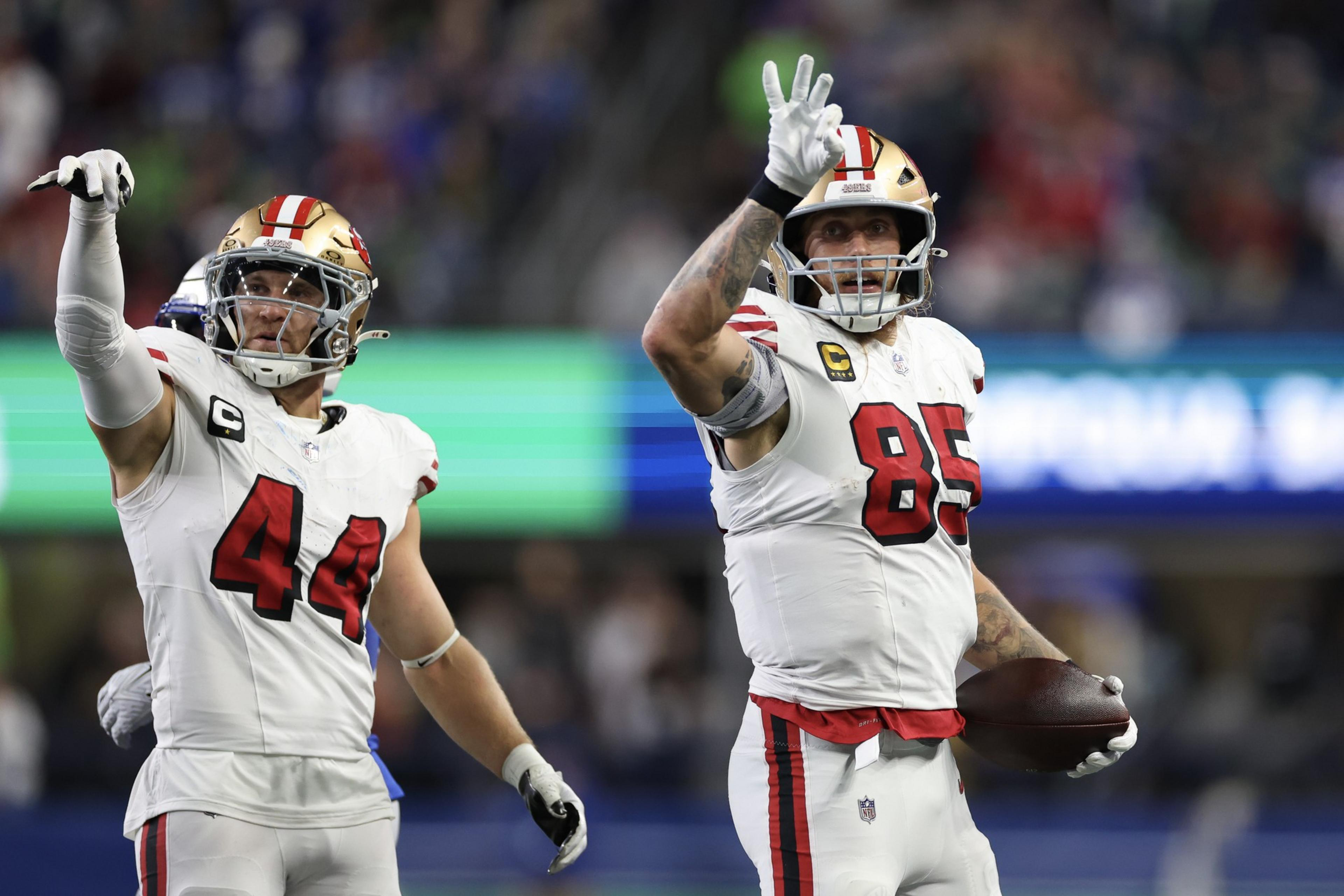 Two football players in white and red uniforms are on a field. One holds a football and gestures with his hand, while the other points outward, both wearing helmets.