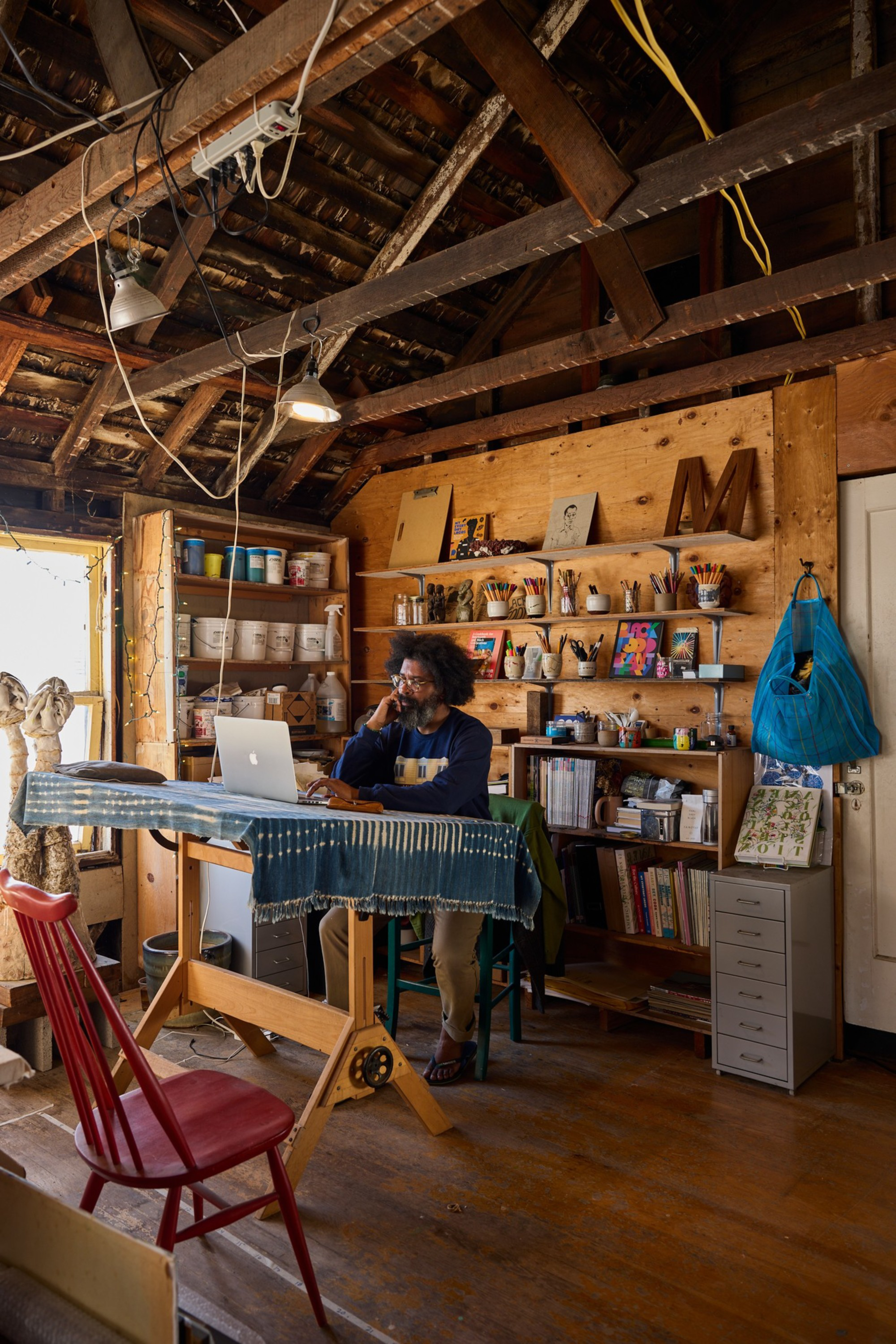A person with curly hair sits at a wooden drafting table covered with a striped cloth, working on a laptop in a cozy, art-filled room.