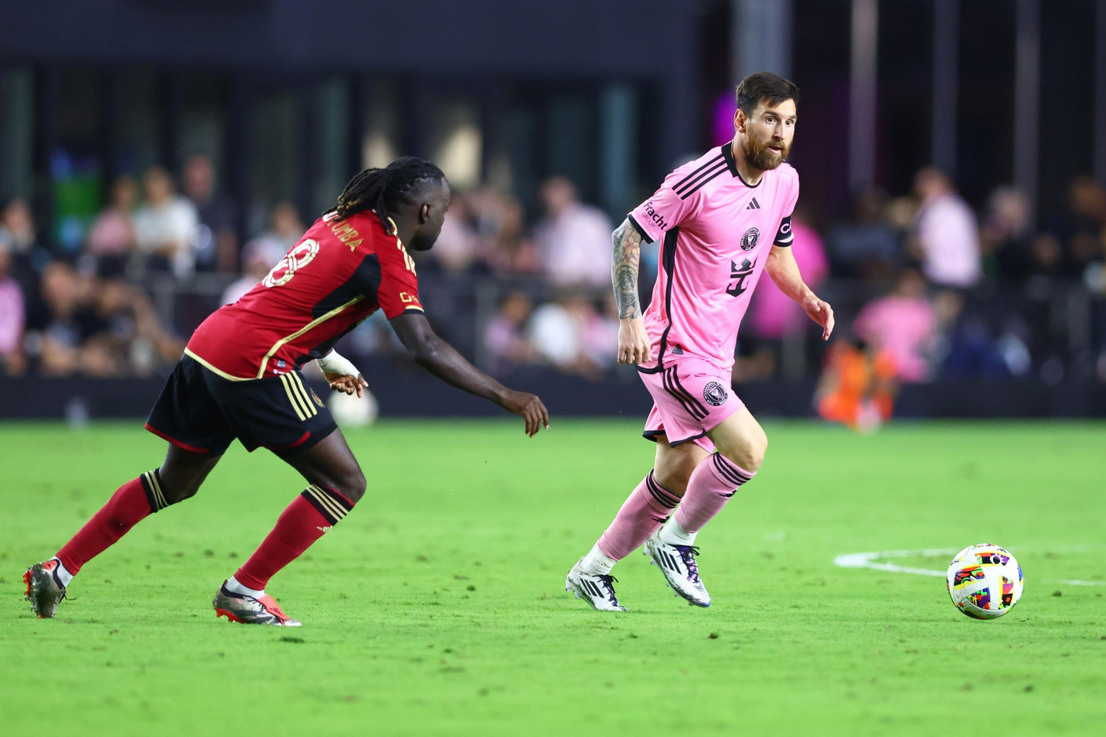 Two soccer players are on a field, one in a pink uniform dribbling the ball, while the other in a red and black uniform pursues him. The background is blurred.