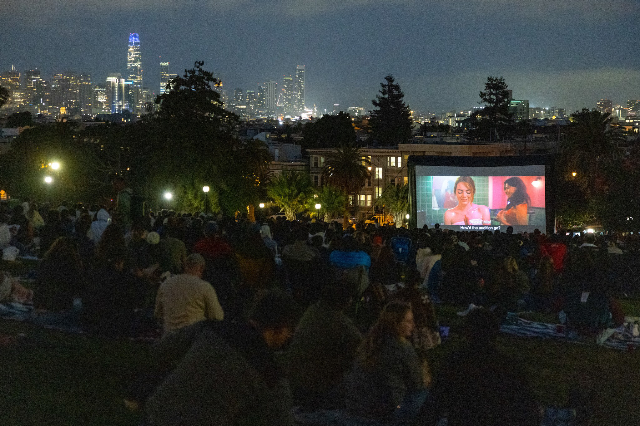 A large crowd sits on a grassy hill watching a movie on an outdoor screen at night, with a city skyline lit up in the background.