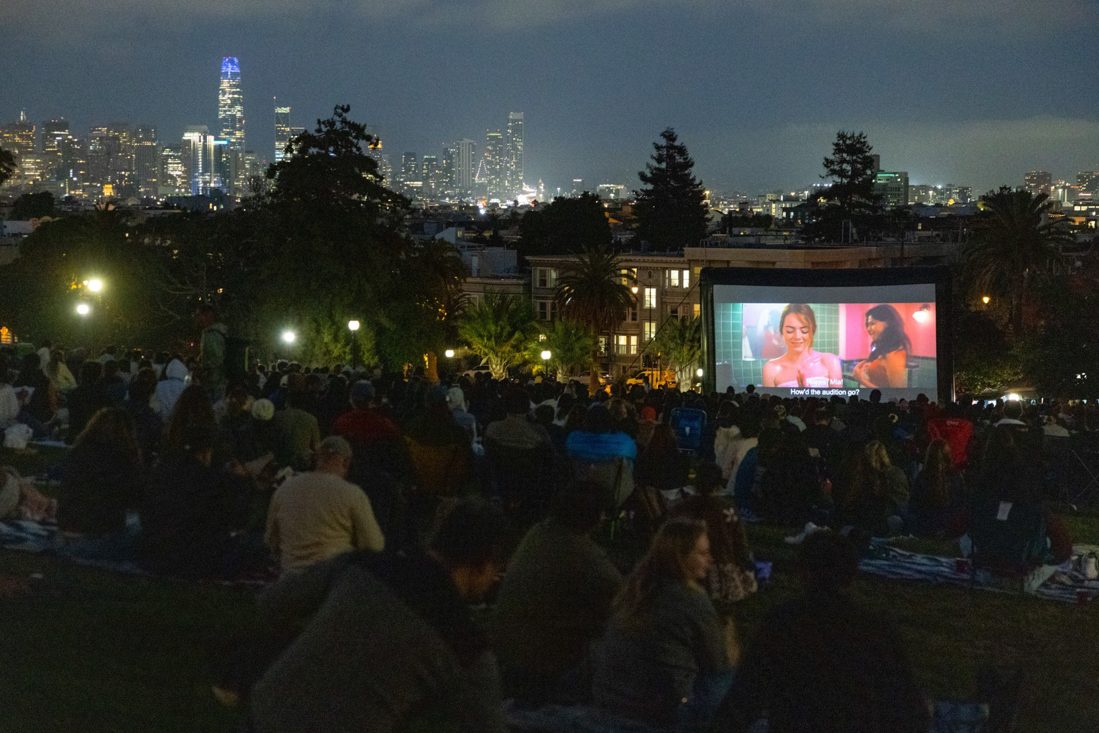A large crowd sits on a grassy hill watching a movie on an outdoor screen at night, with a city skyline lit up in the background.