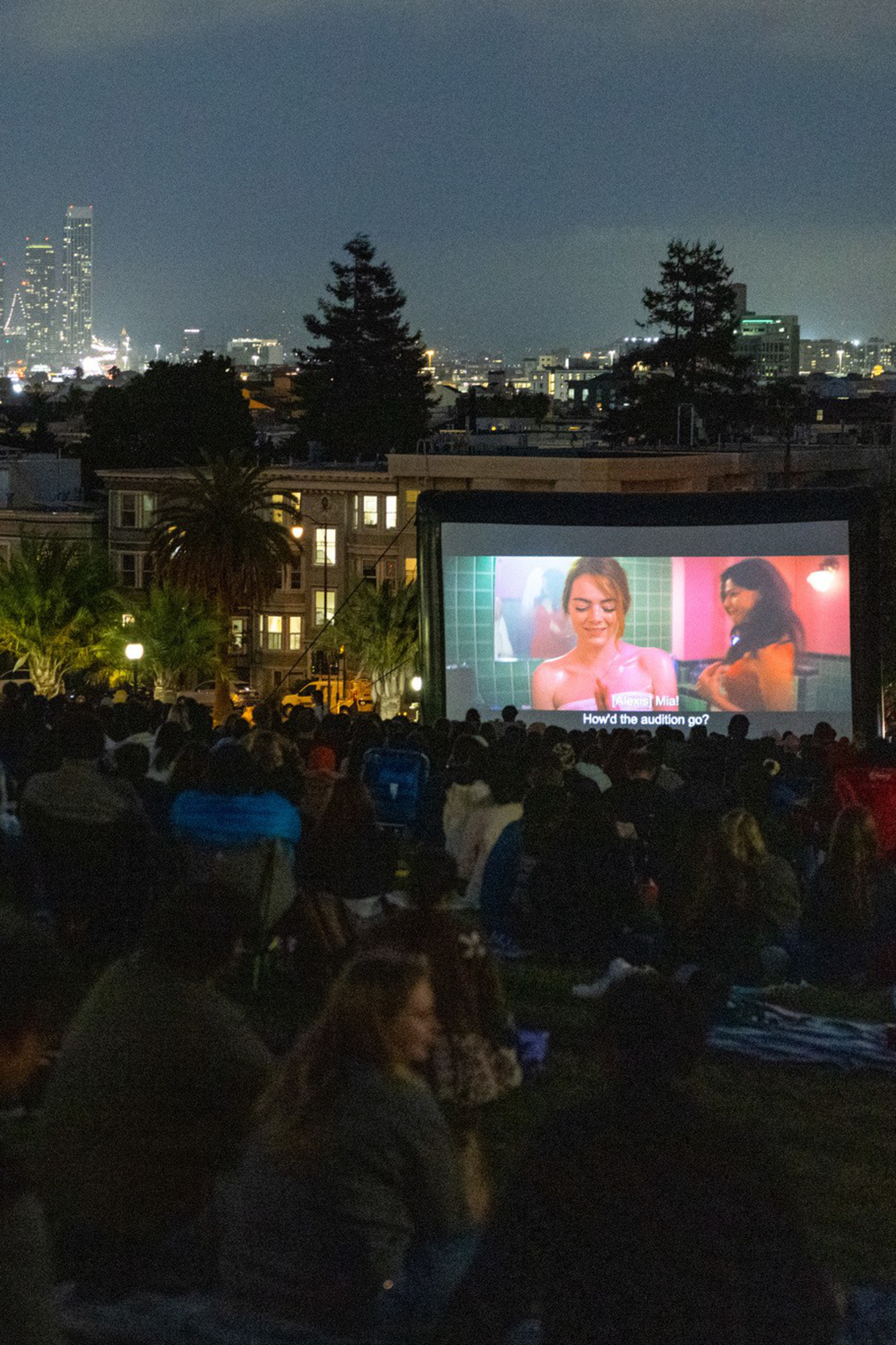 A large crowd sits on a grassy hill watching a movie on an outdoor screen at night, with a city skyline lit up in the background.