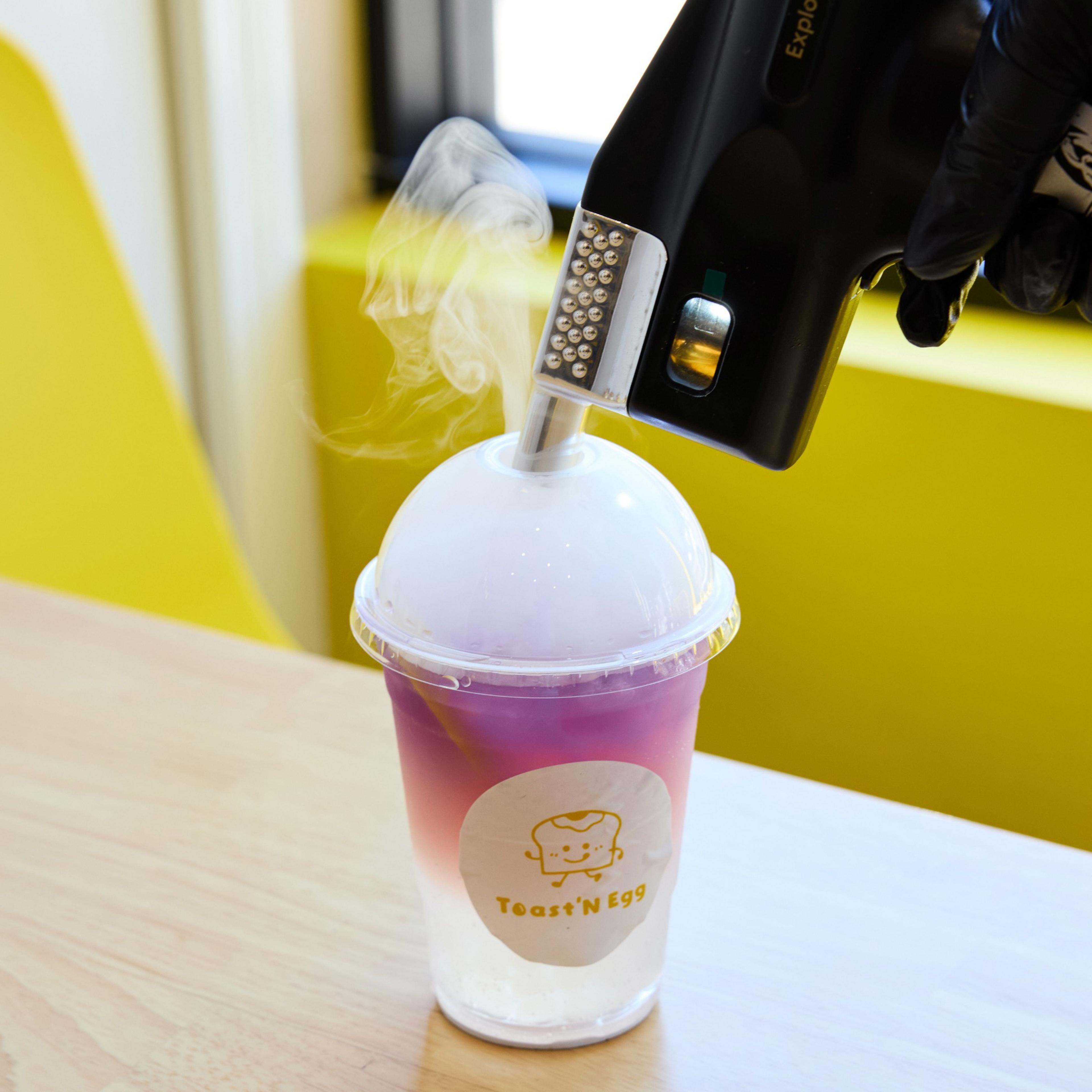A gloved hand holds a device emitting vapor into a dome-lidded, purple gradient drink at a table, labeled &quot;Toast'n Egg,&quot; with yellow background elements.