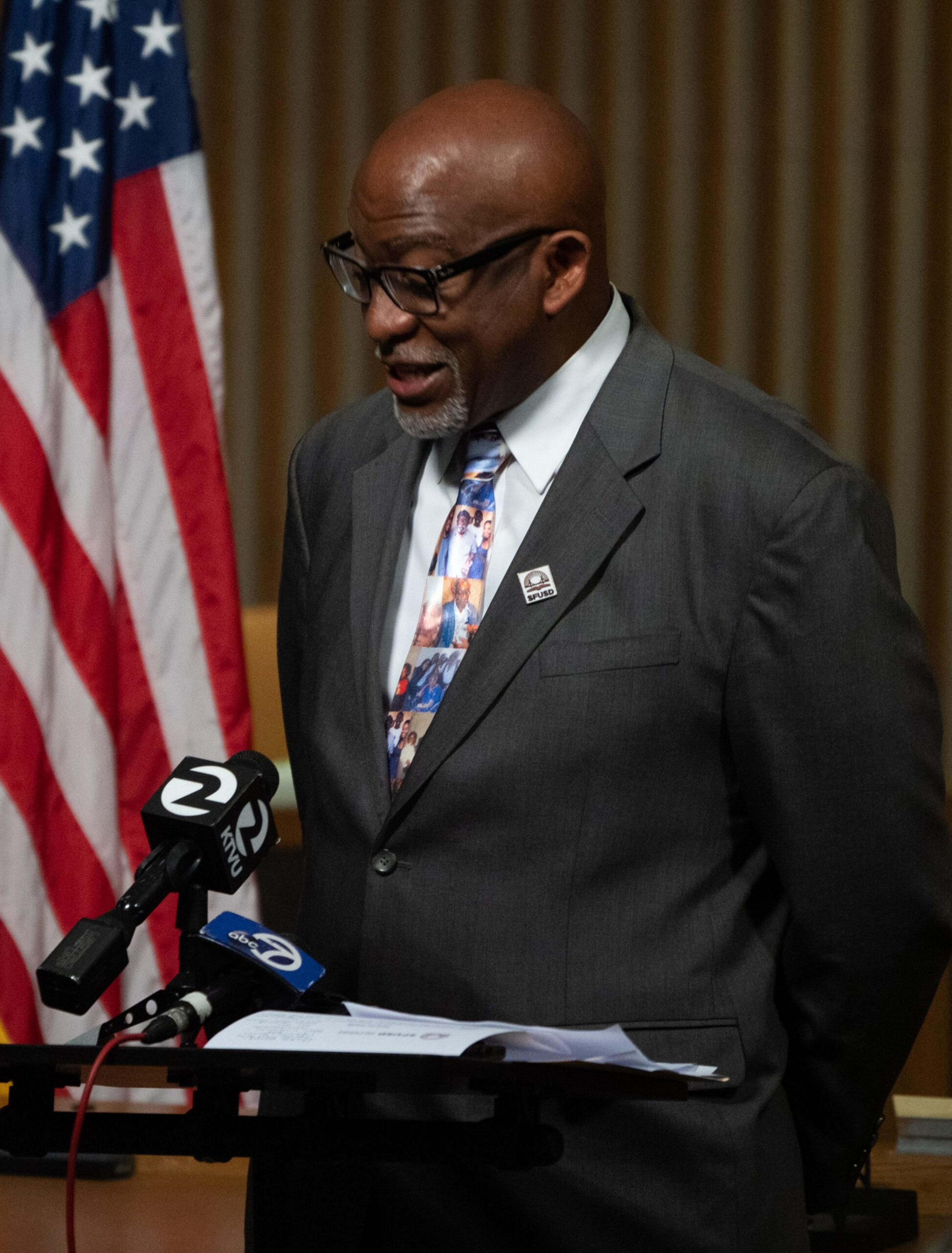 A man in a gray suit and patterned tie stands at a podium with microphones, speaking, with an American flag visible behind him.