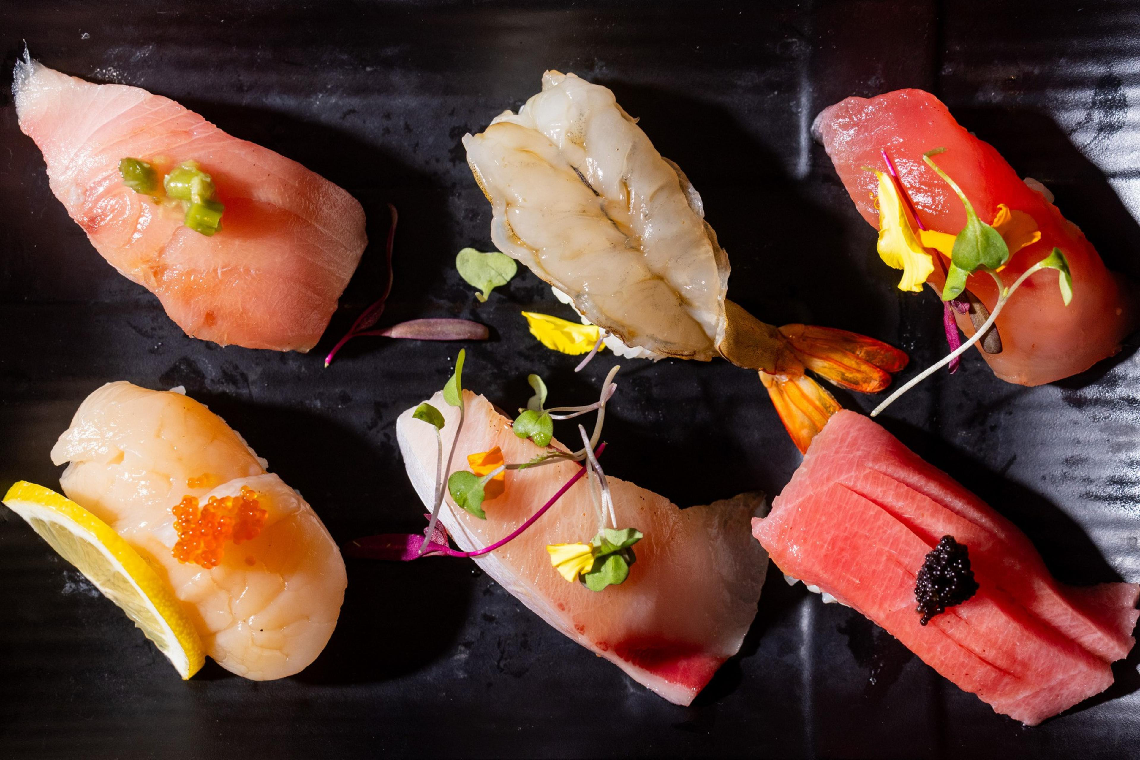 The image shows a variety of beautifully arranged sushi on a black plate, with garnishes like microgreens and a lemon slice, highlighting the fresh seafood toppings.