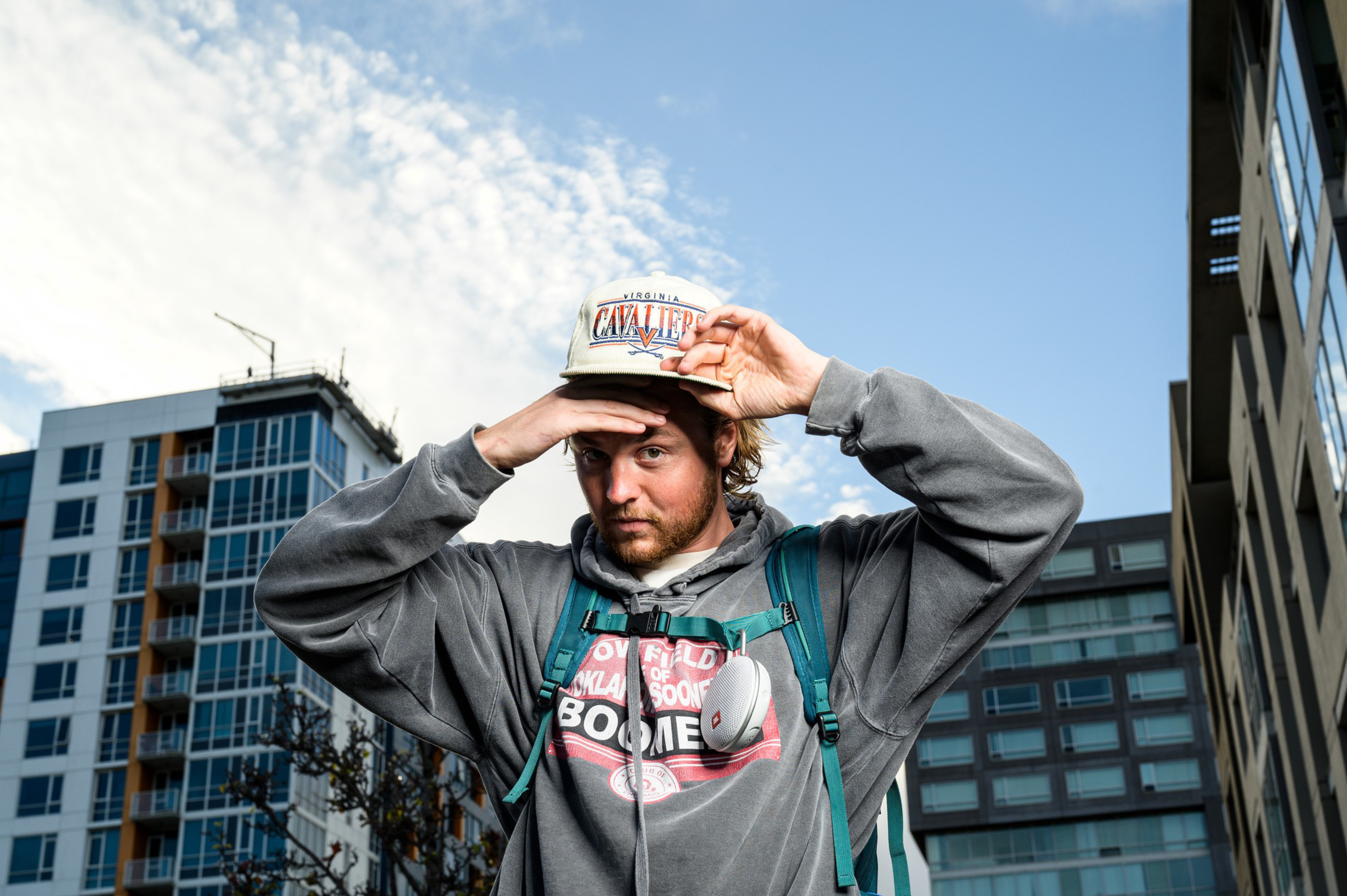 A man wearing a gray hoodie and a Virginia Cavaliers cap adjusts his hat while standing between modern apartment buildings under a partly cloudy sky.