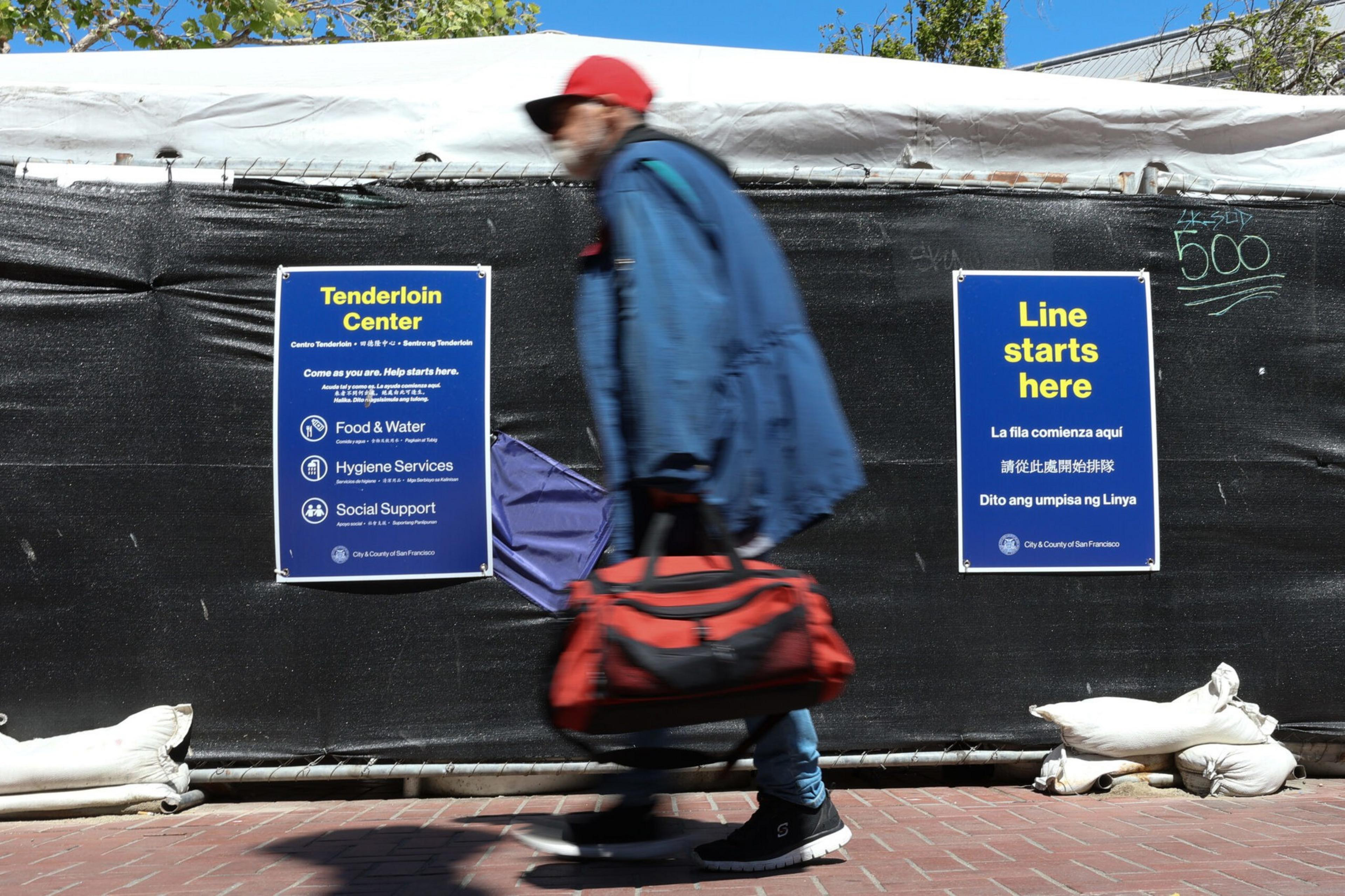 A person wearing a red cap and blue jacket walks past black barriers with signs detailing services at the Tenderloin Center and where the line starts.