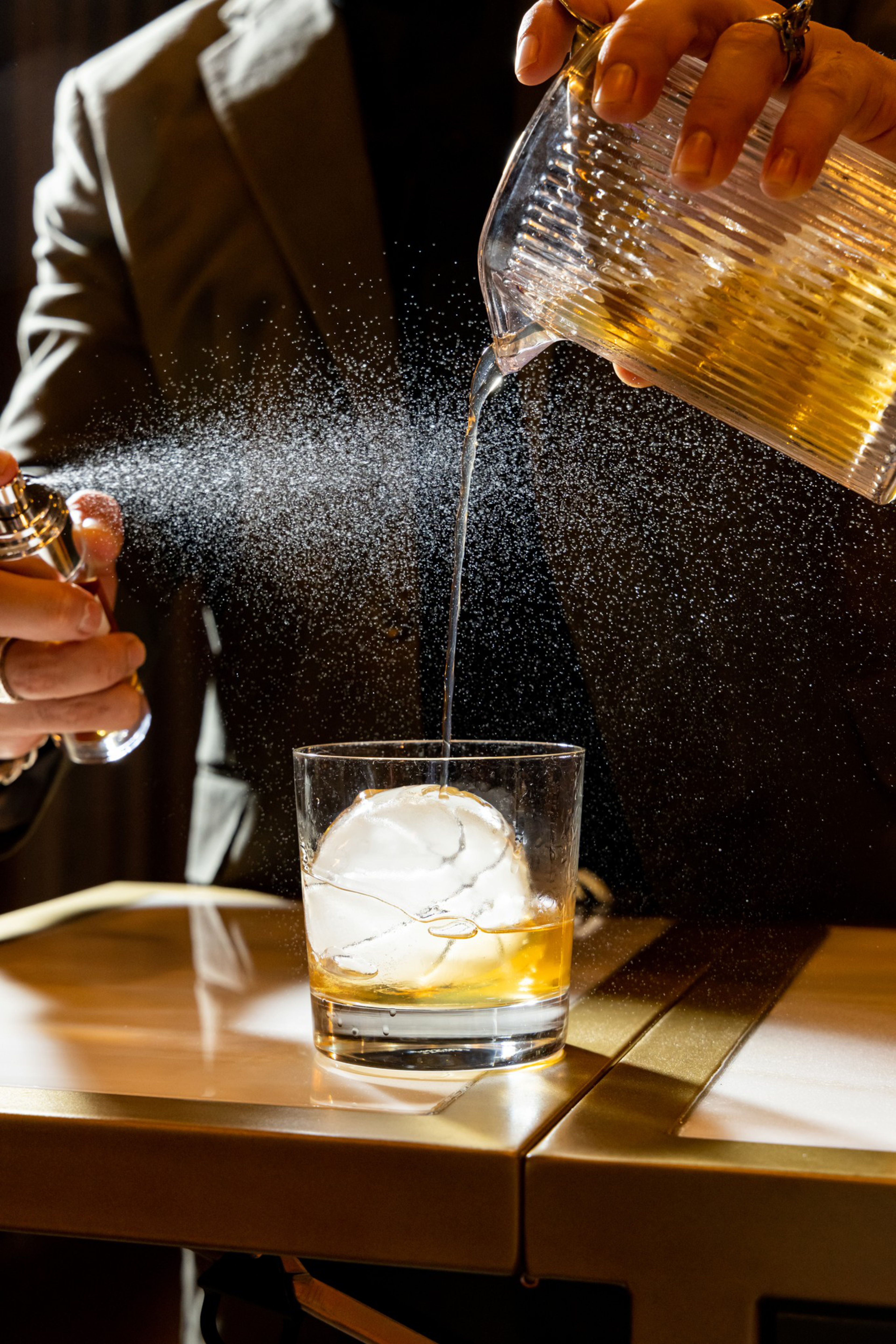 A person pours a drink over a large ice sphere in a glass while spraying a fine mist nearby, with light reflecting on the table.