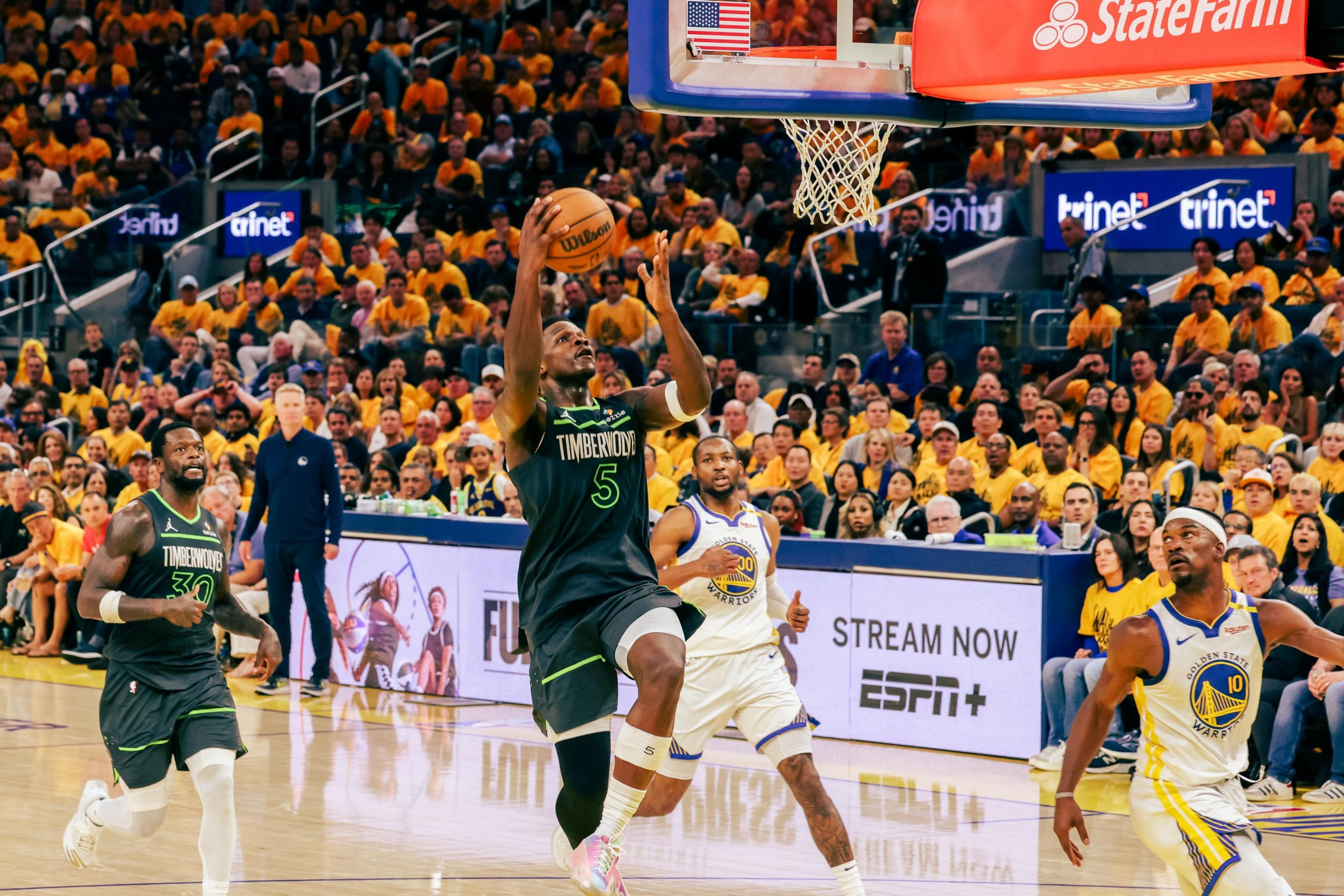 A basketball player in a Timberwolves jersey leaps toward the hoop, while two Warriors players watch. The crowd in yellow is engaged in the background.