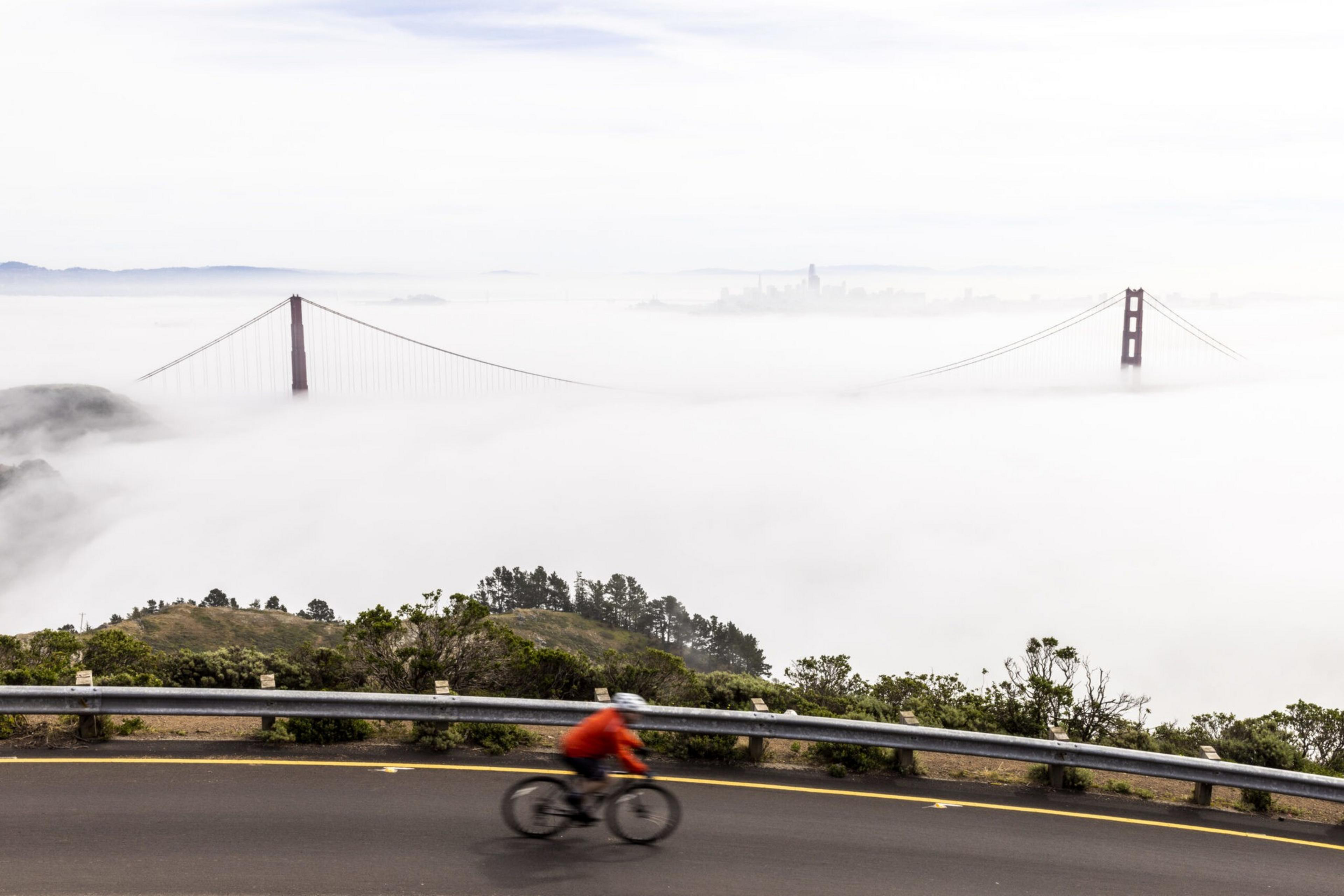 A cyclist in an orange jacket rides a road curving along a hillside, with the Golden Gate Bridge towers partially visible above thick fog.