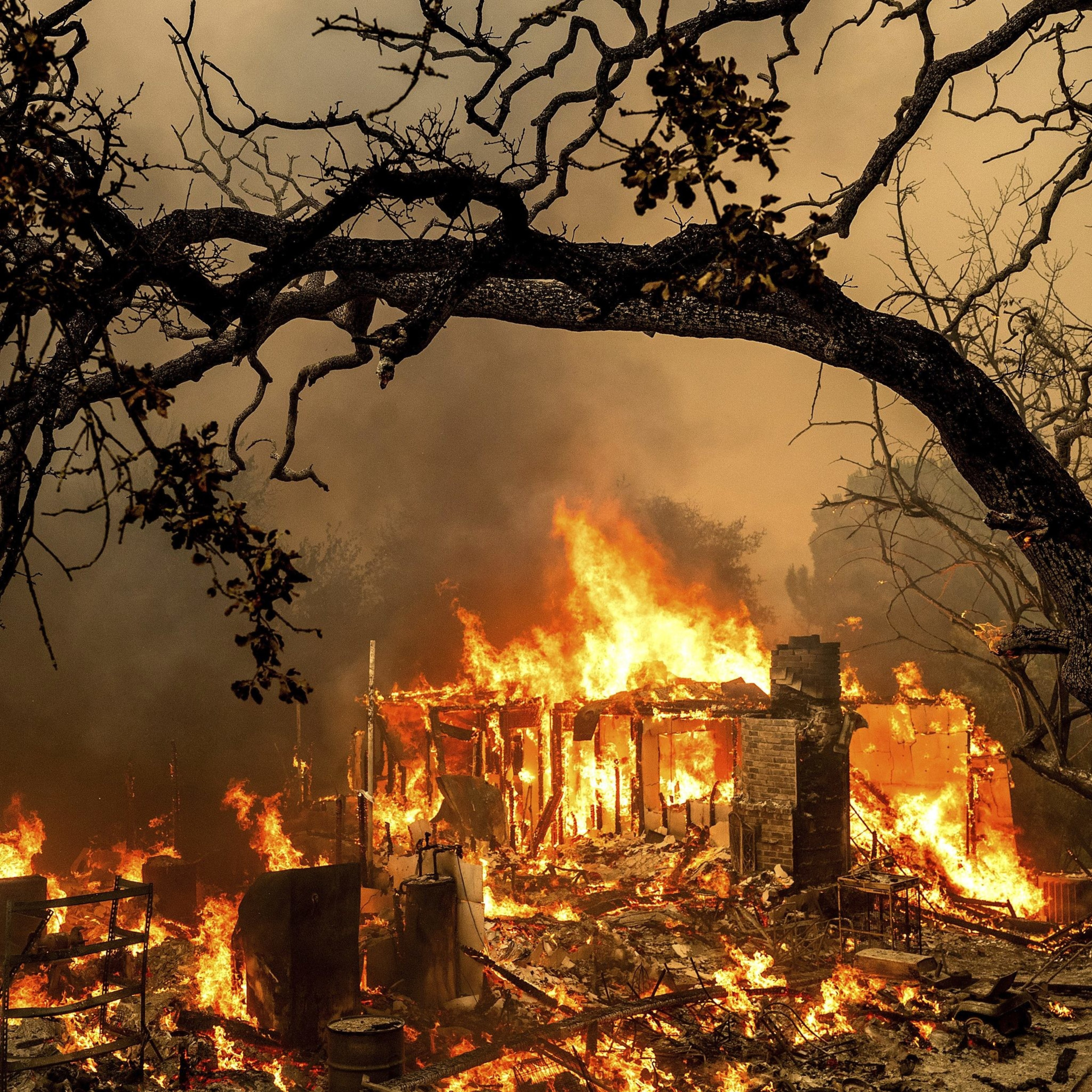 A house is engulfed in intense flames, with bright orange fire consuming its structure. Dark smoke billows, and silhouetted trees frame the scene.