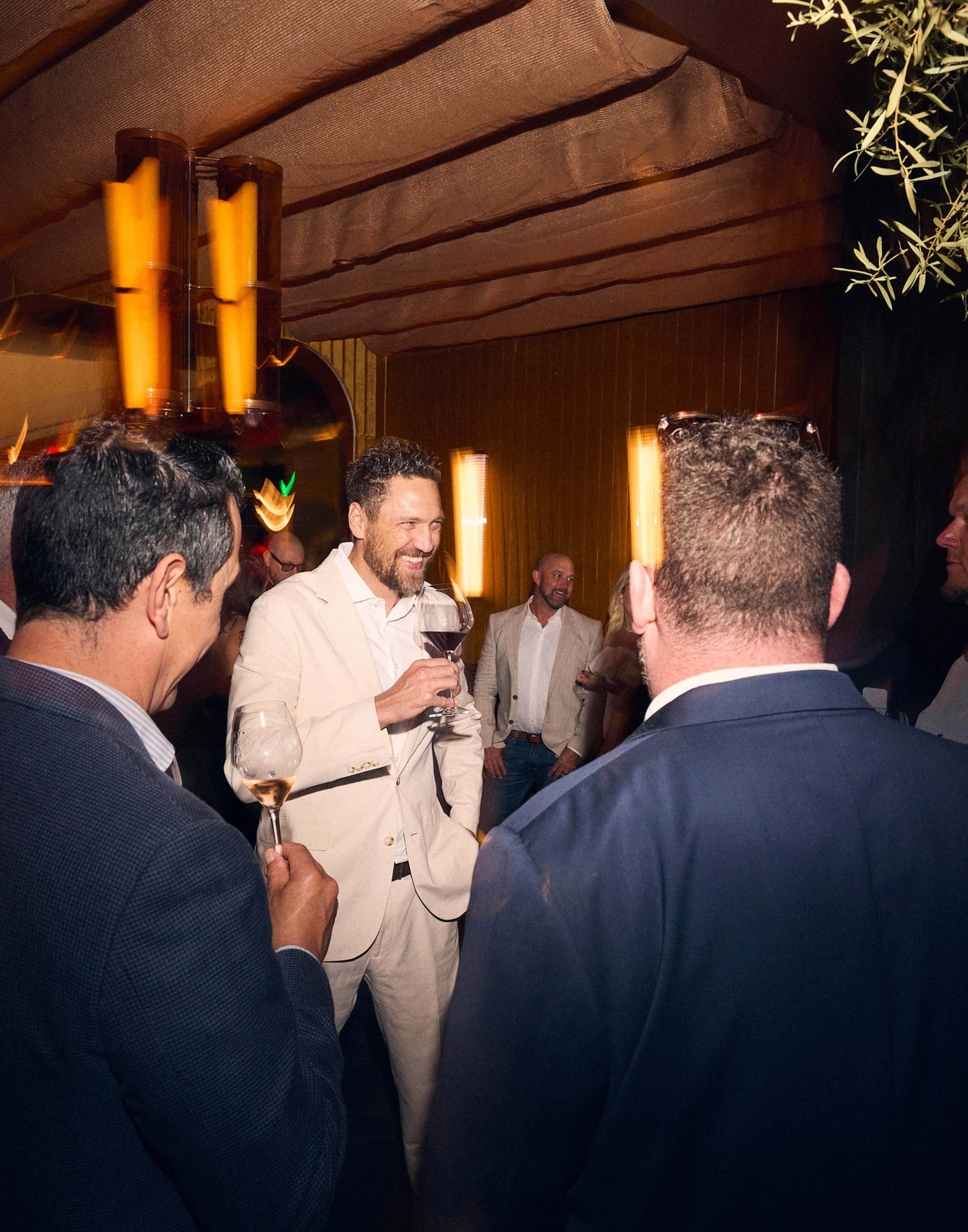 A group of men in suits socialize indoors, holding wine glasses and smiling under warm, dim lighting with yellow walls and fabric ceiling panels.