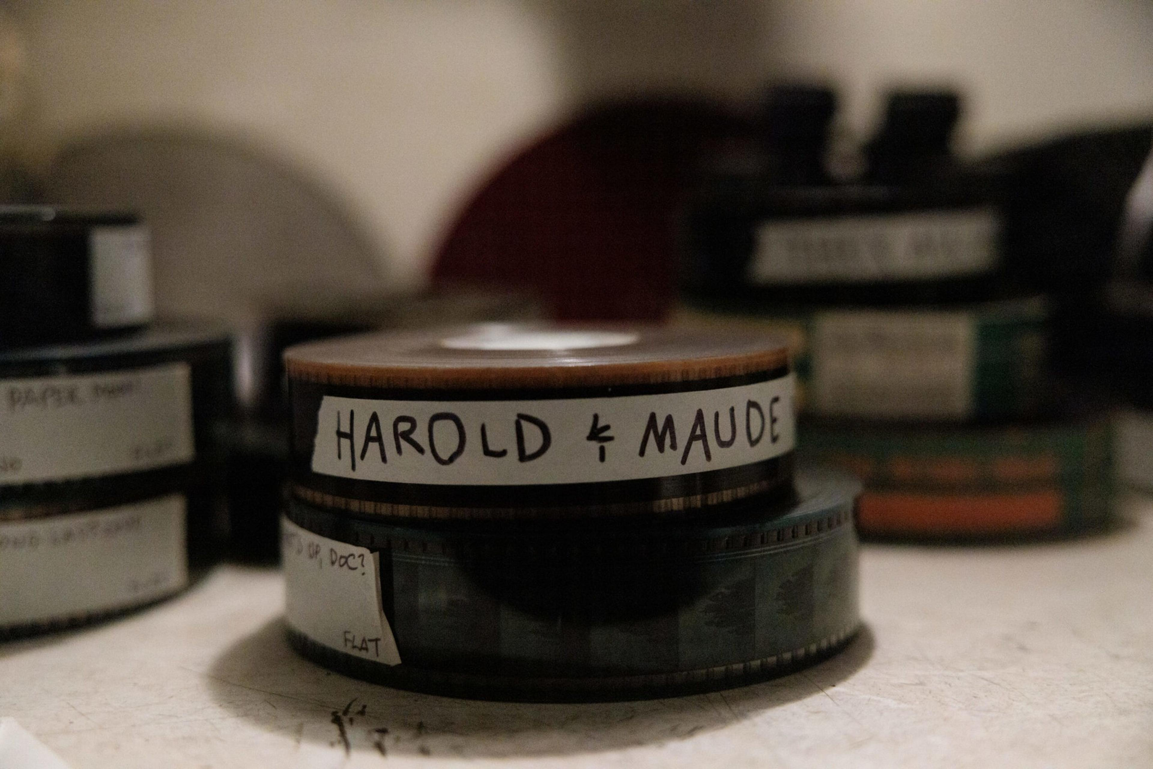 Two film reels stacked on each other, the top one labeled "HAROLD & MAUDE," placed on a surface with other blurred film reels in the background.