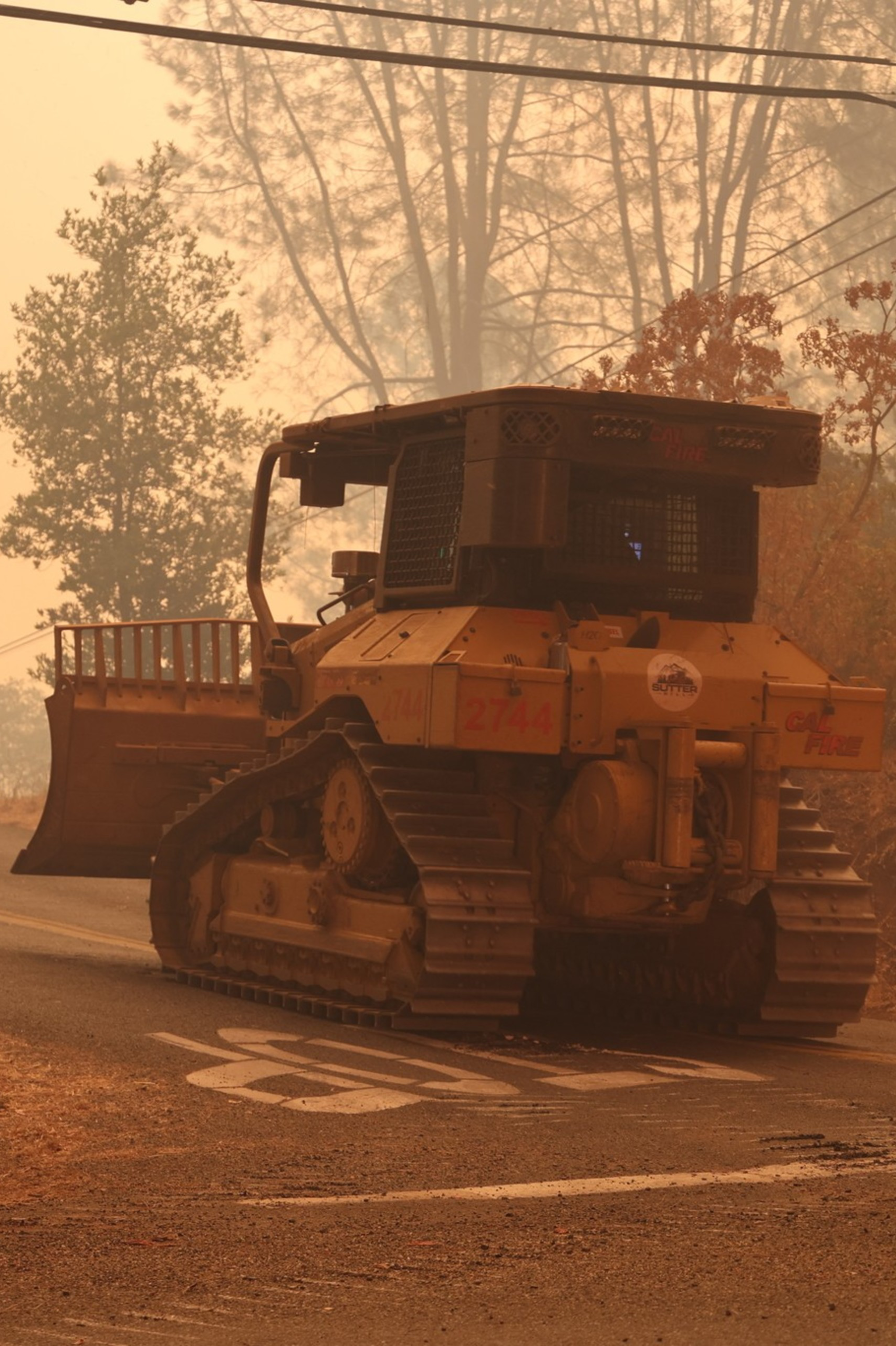 Two large bulldozers are on a smoky, rural road, near a signpost reading &quot;Cohasset Rd&quot; and a &quot;For Sale&quot; sign. The area is hazy, likely due to wildfire conditions.