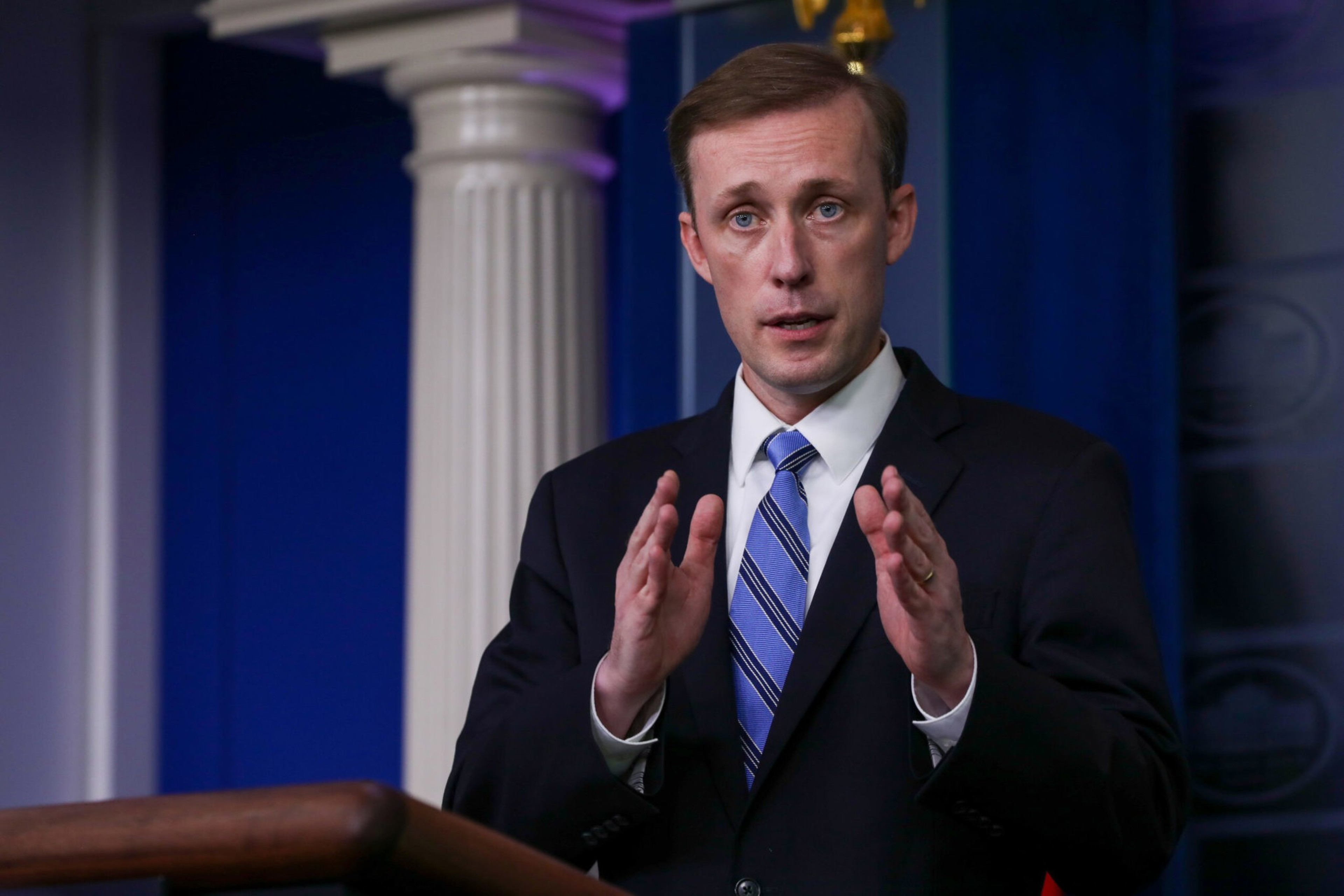 A man in a dark suit and striped blue tie gestures with both hands while speaking at a podium in front of blue and white columns.
