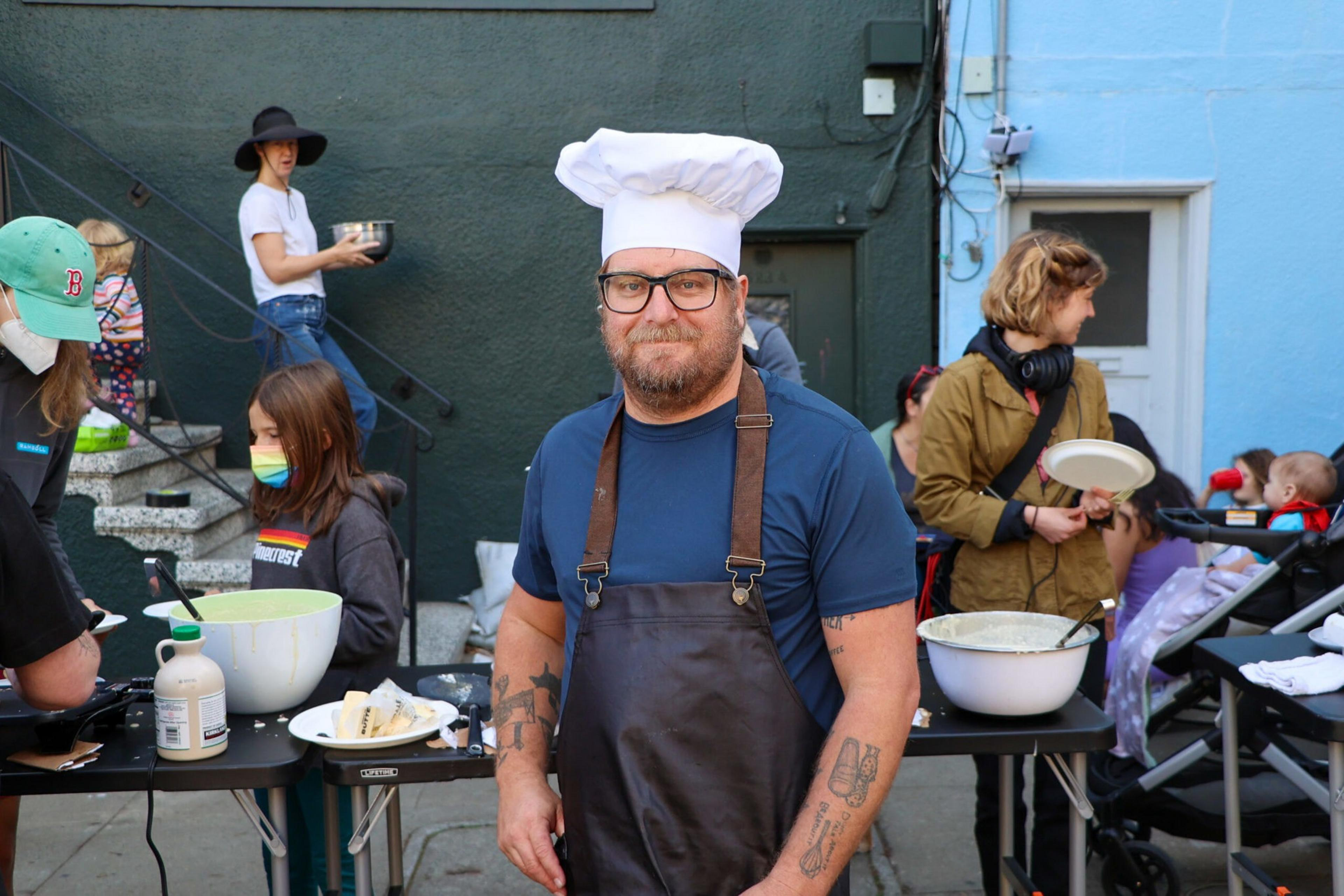 A man in a chef's hat and apron stands smiling outdoors, surrounded by people. There's cooking equipment and bowls on tables, and others are holding plates.