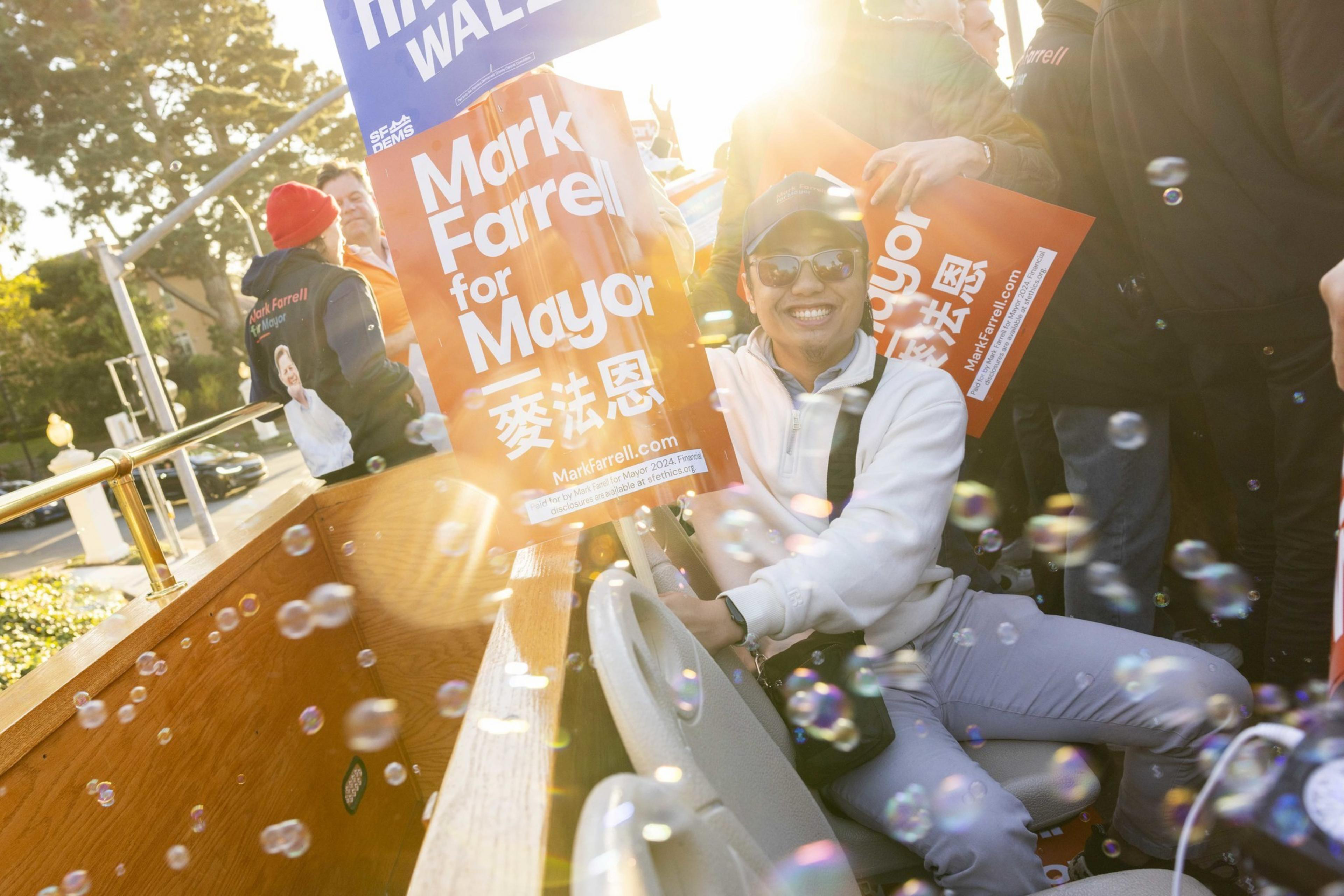 A smiling person in sunglasses holds a &quot;Mark Farrell for Mayor&quot; sign while sitting on a vehicle surrounded by sunlight and soap bubbles.
