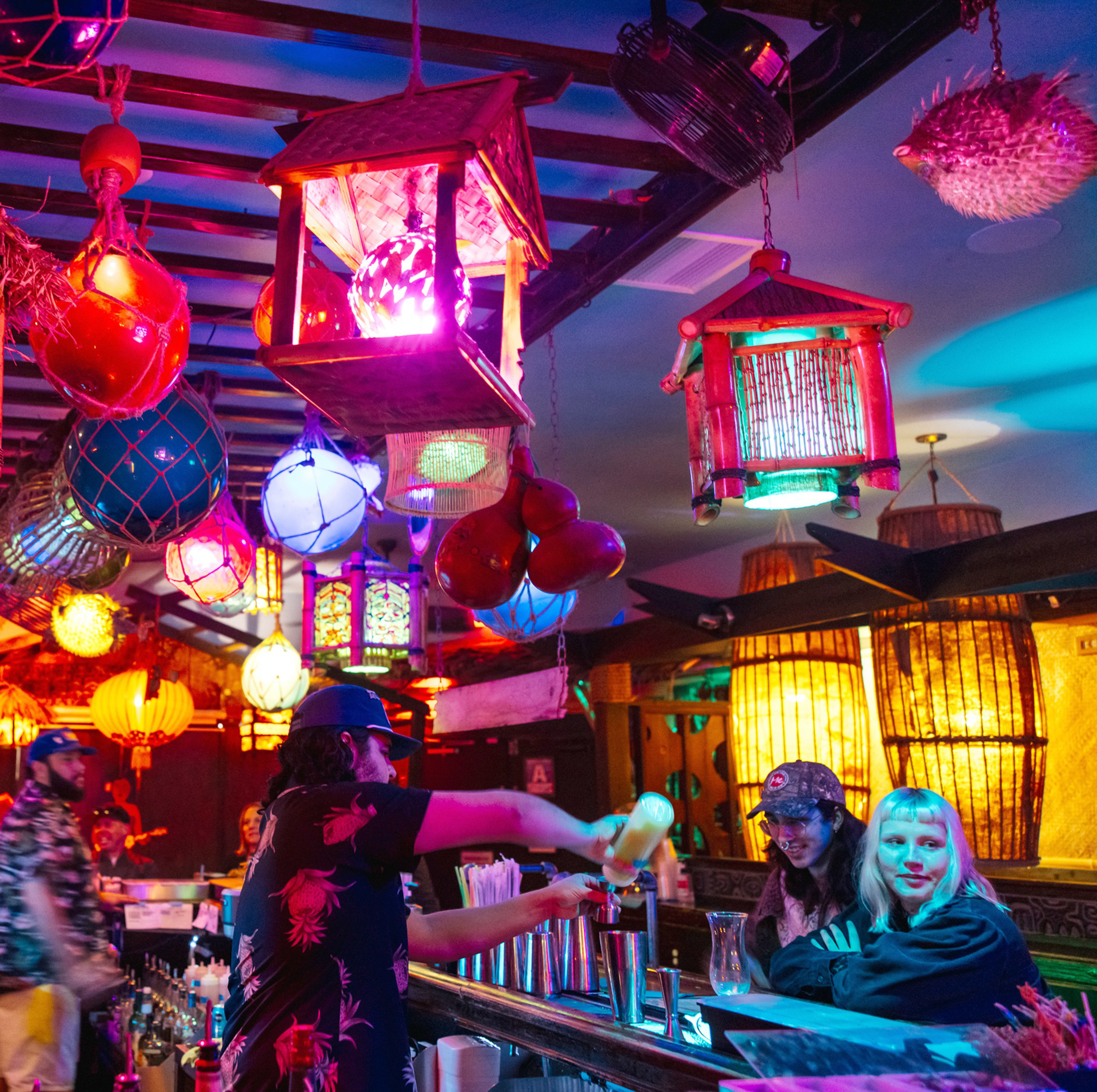 A colorful bar scene features vibrant lanterns hanging from the ceiling, a bartender preparing drinks, and patrons chatting at the counter under warm, multicolored lights.