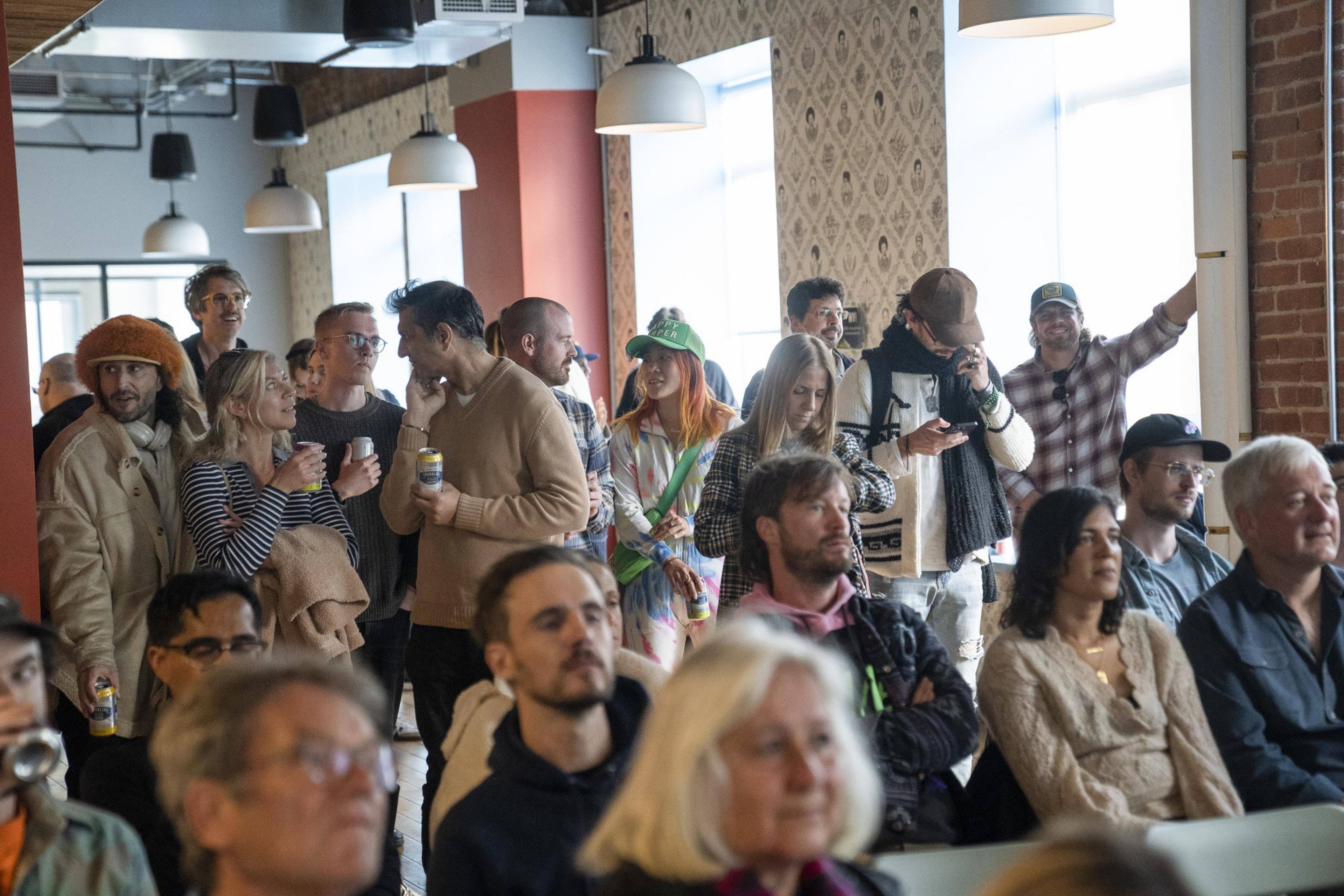 A group of people gathers in a well-lit room, some standing and chatting with drinks, others seated attentively, all under modern hanging lights.